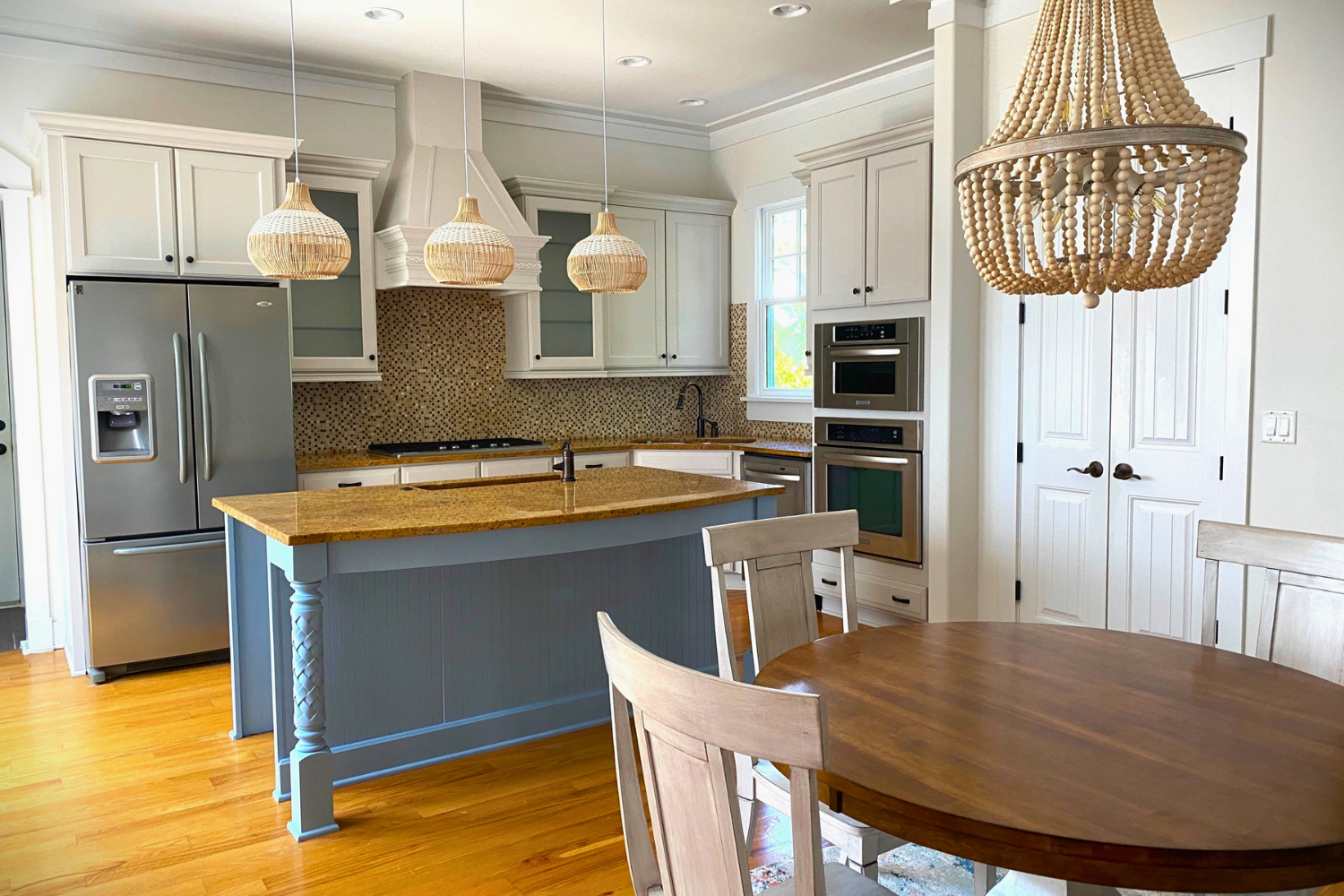 Kitchen with white cabinets, a yellow granite island, stainless steel appliances, a wooden dining table with chairs, and hanging woven light fixtures.