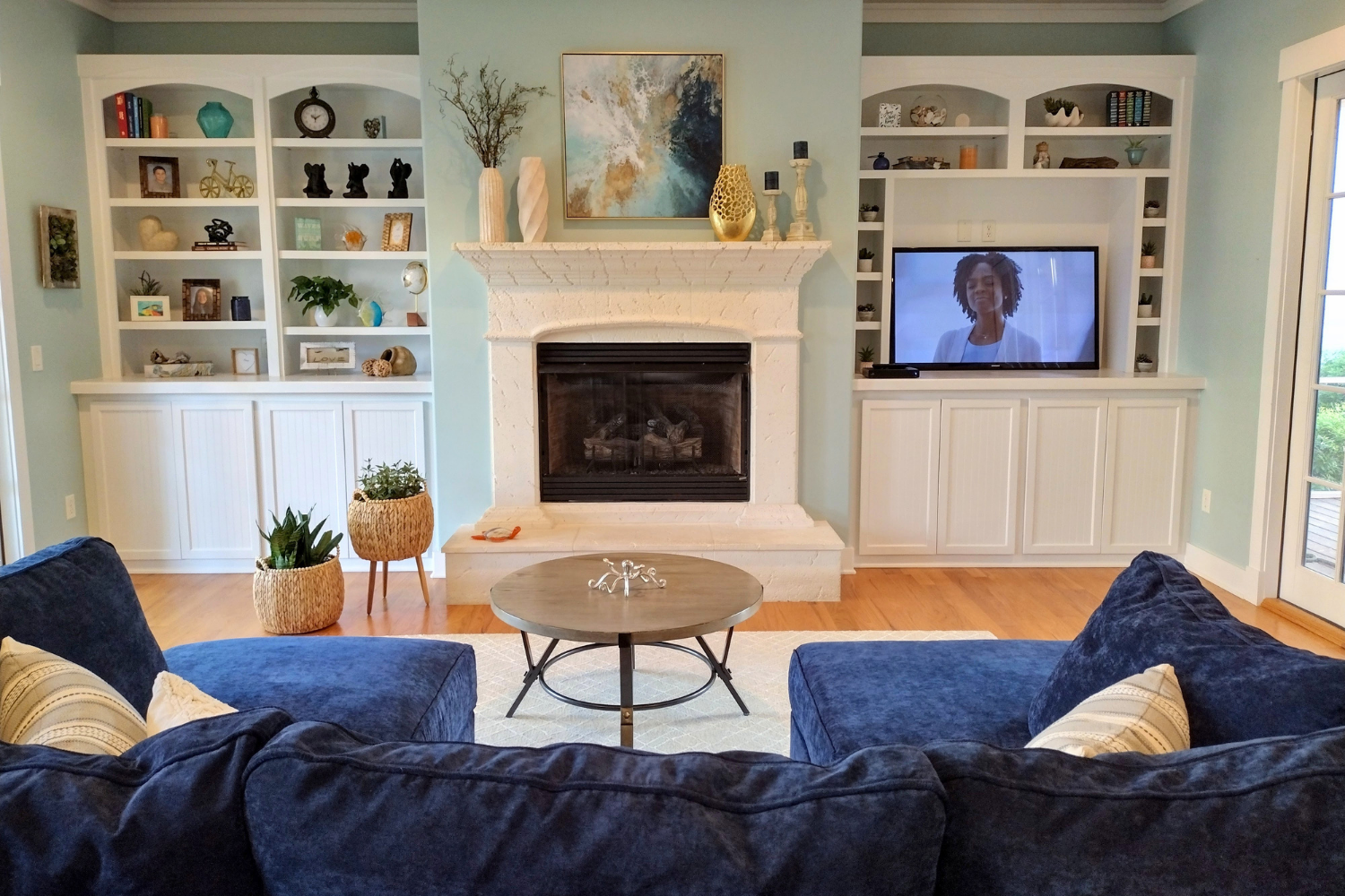 Living room with a blue sofa, round coffee table, white fireplace mantel, and built-in shelves with decorative items and books. A TV is on the right shelf, and a framed painting hangs above the fireplace. Two potted plants are on the floor to the lef