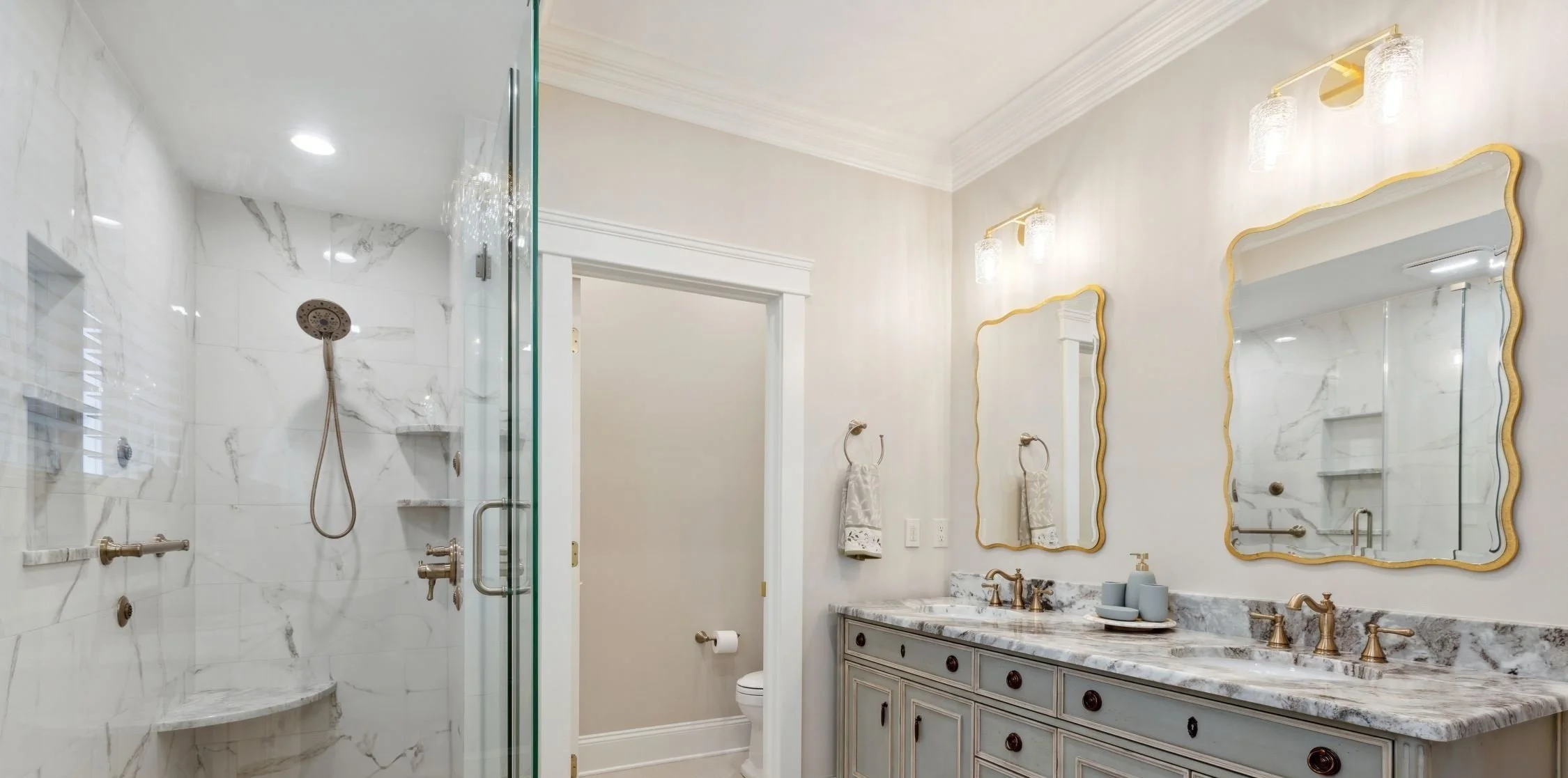 Elegant bathroom with marble-tiled glass shower, double vanity, gold-framed mirrors, and crystal lighting