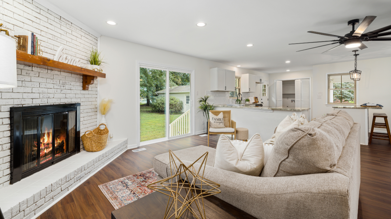 Living room with a fireplace, white brick wall, beige sofa, pillows, a gold decorative star, a colorful rug, sliding glass door leading outside, and an open kitchen with white cabinets and a breakfast bar.