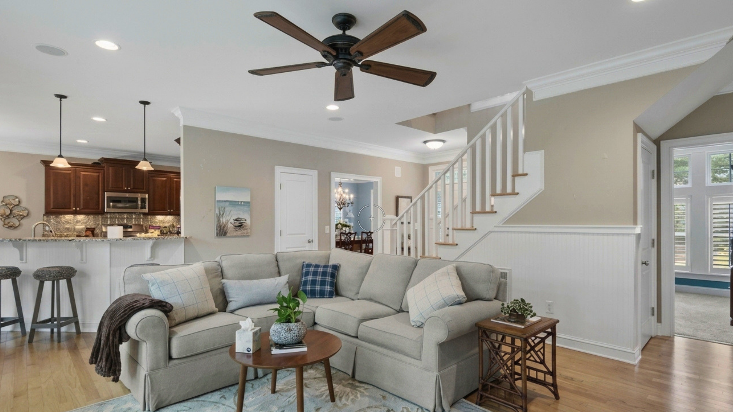 Living room with beige sofa, throw pillows, small wooden coffee table, kitchen area with pendant lights, dark wood cabinets, granite countertops, staircase with white railings, and large windows.