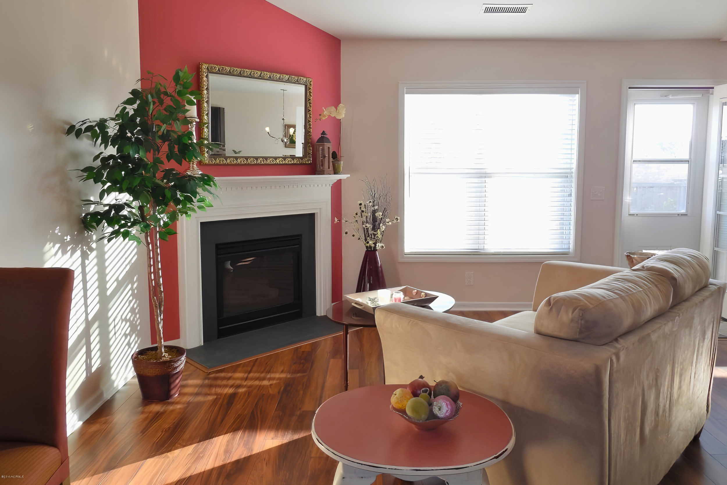 Living room with a white sofa, a pink coffee table with decorative balls, a tall potted plant, a fireplace with a gold-framed mirror above it, a large window with blinds, and a door leading outside.