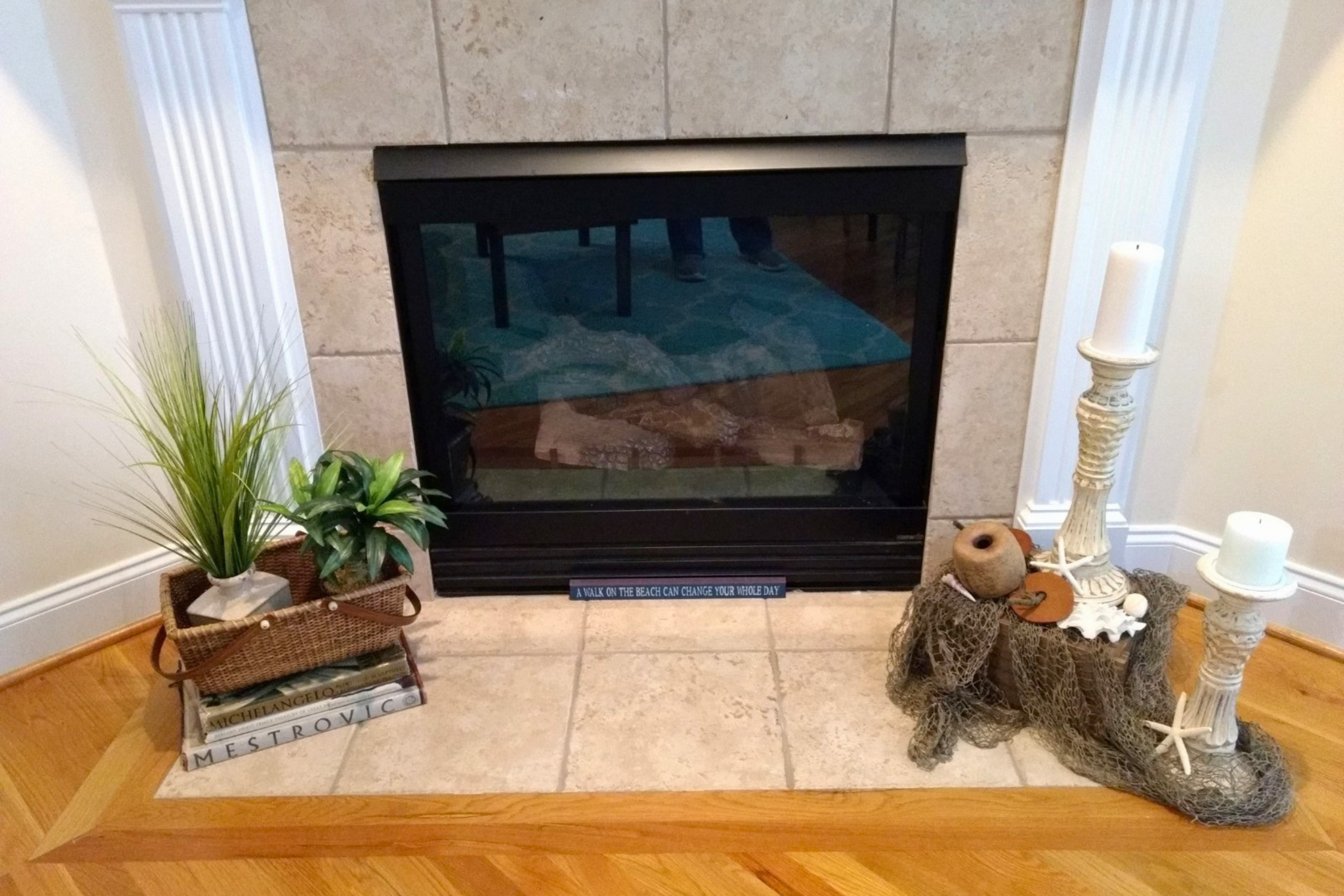 A fireplace with a tile surround and a sign that says "A walk on the beach can change your whole day." There are decorative items to the right including a white candlestick holder with two candles, a ceramic vase, and a basket with dried seaweed or coral. To the left, there are green indoor plants and a stack of books with titles related to Michelangelo and architecture on a small wooden table.