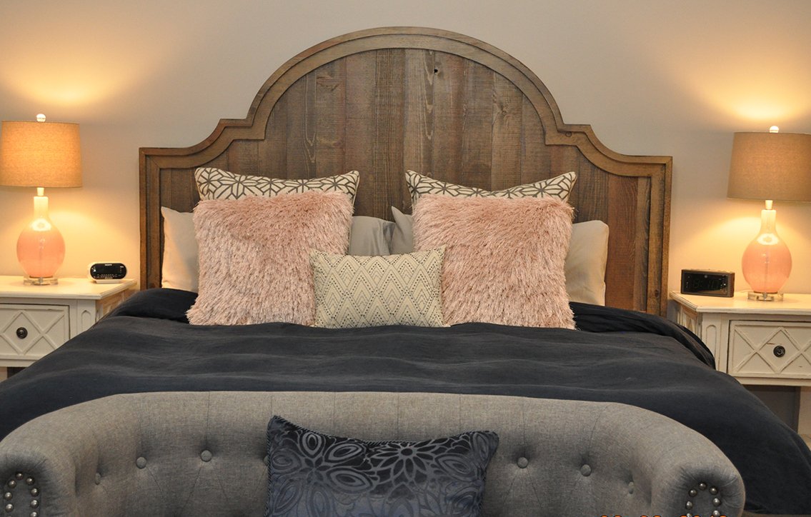 A neatly made bed with a wooden headboard, pink fluffy pillows, patterned pillows, and a black bedspread. Flanking the bed are cream-colored nightstands with pink lamps and small clocks, and a tufted bench with a decorative pillow at the foot of the 