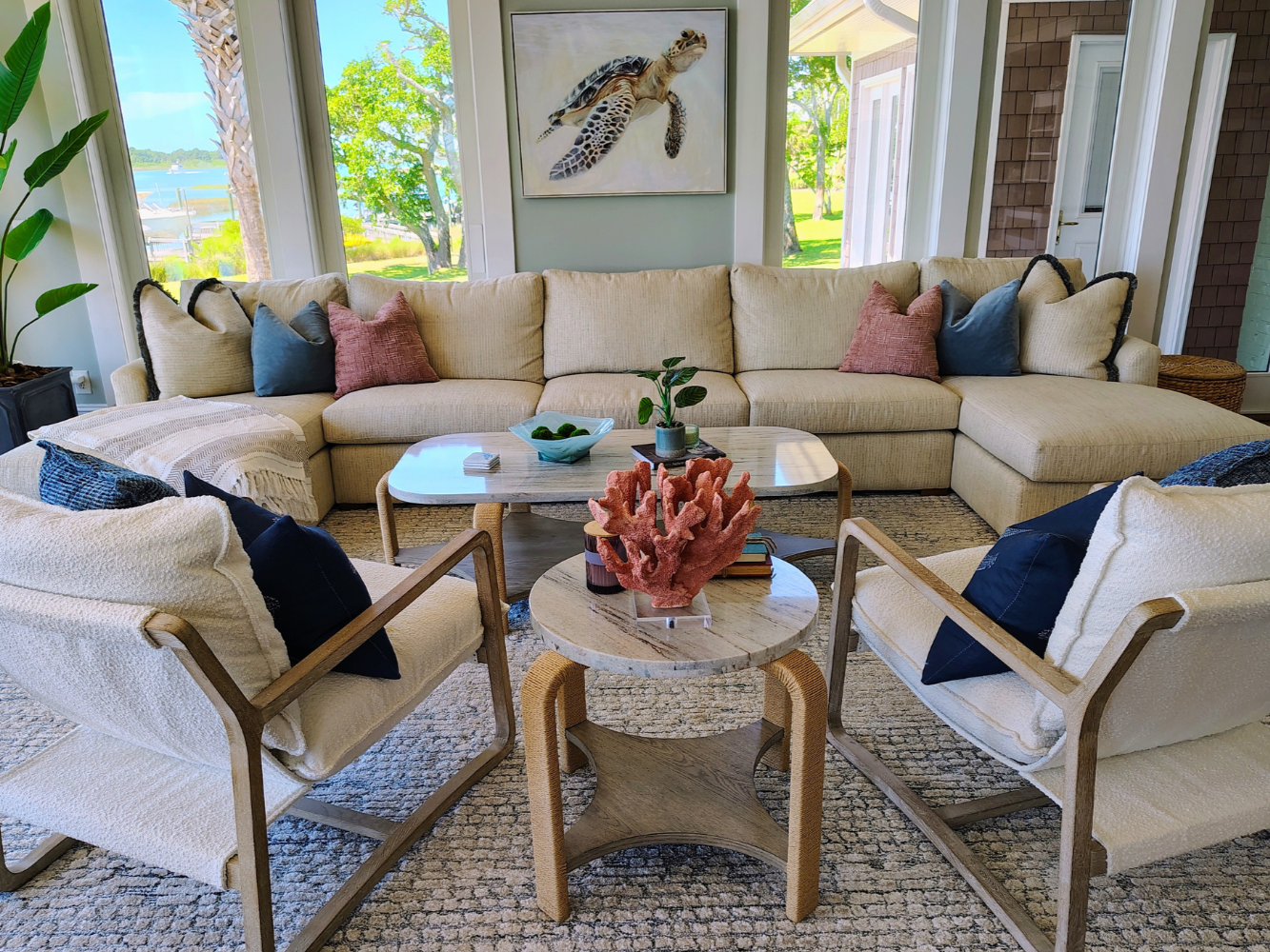 Living room with large sofa, two armchairs, coffee tables, plants, colorful cushions, and a turtle artwork on the wall, overlooking a green outdoor landscape.