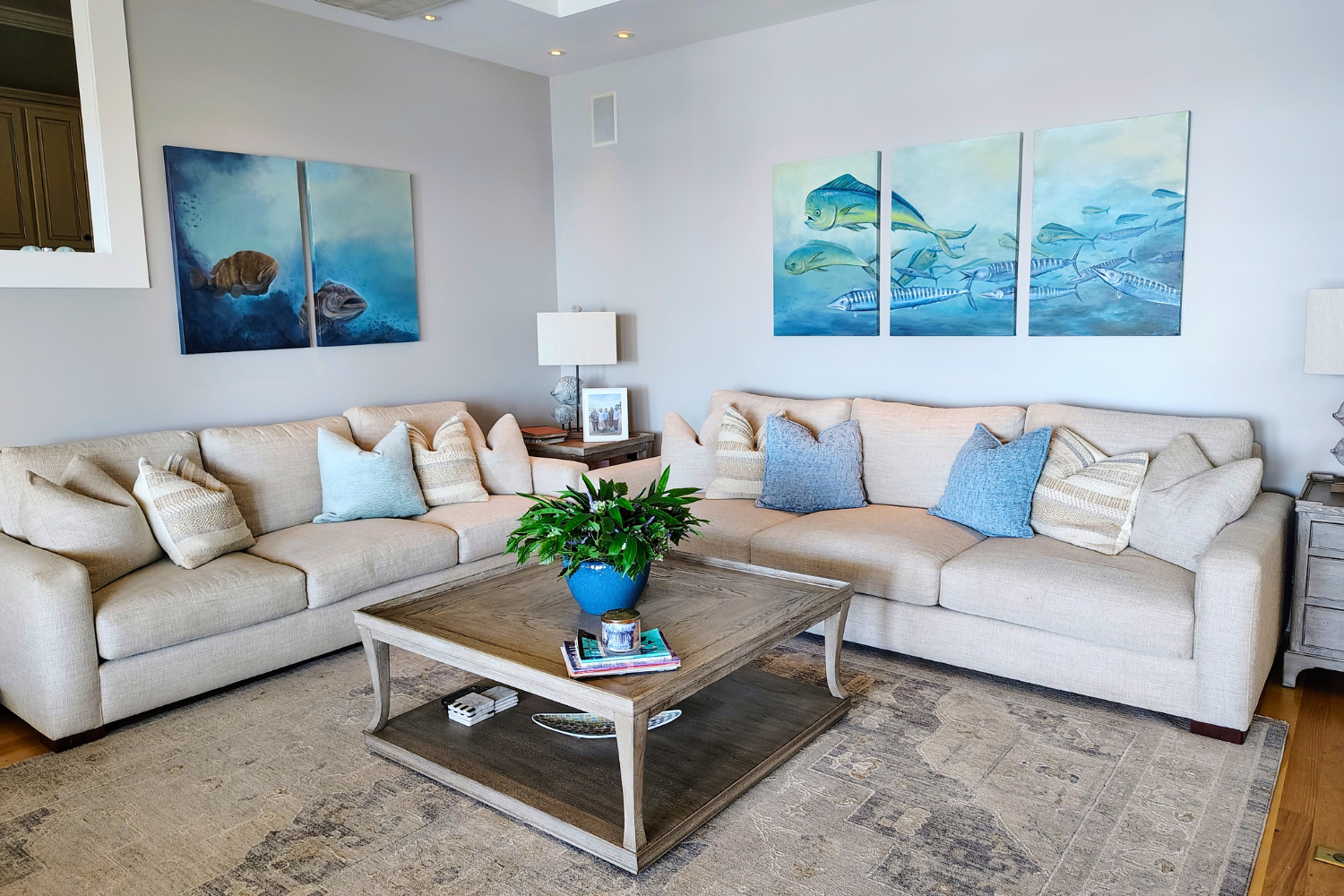 Living room with beige couches, blue and beige throw pillows, a wooden coffee table with a potted green plant, a table lamp, framed photographs, and underwater-themed artwork featuring fish on the walls.
