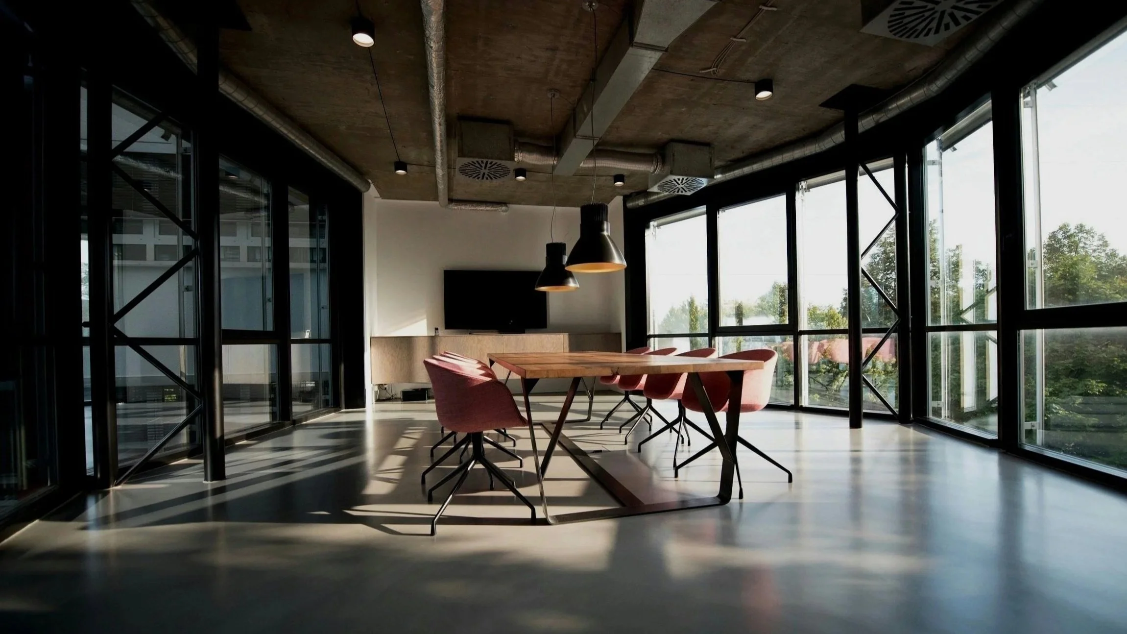 Modern conference room with large windows, a wooden table, pink chairs, black pendant lights, and a television, with sunlight streaming through the windows.