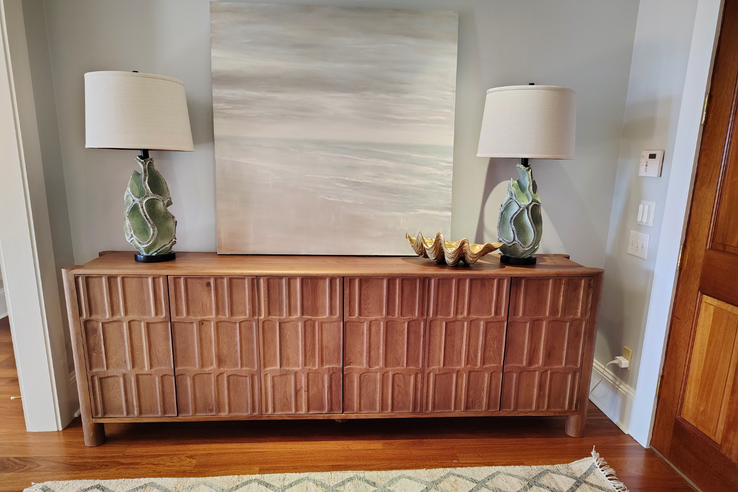 A wooden sideboard with a geometric pattern on its doors, topped with two matching table lamps featuring green and beige decorative bases and beige lampshades, a large abstract painting in neutral tones, and a decorative shell sculpture across from a