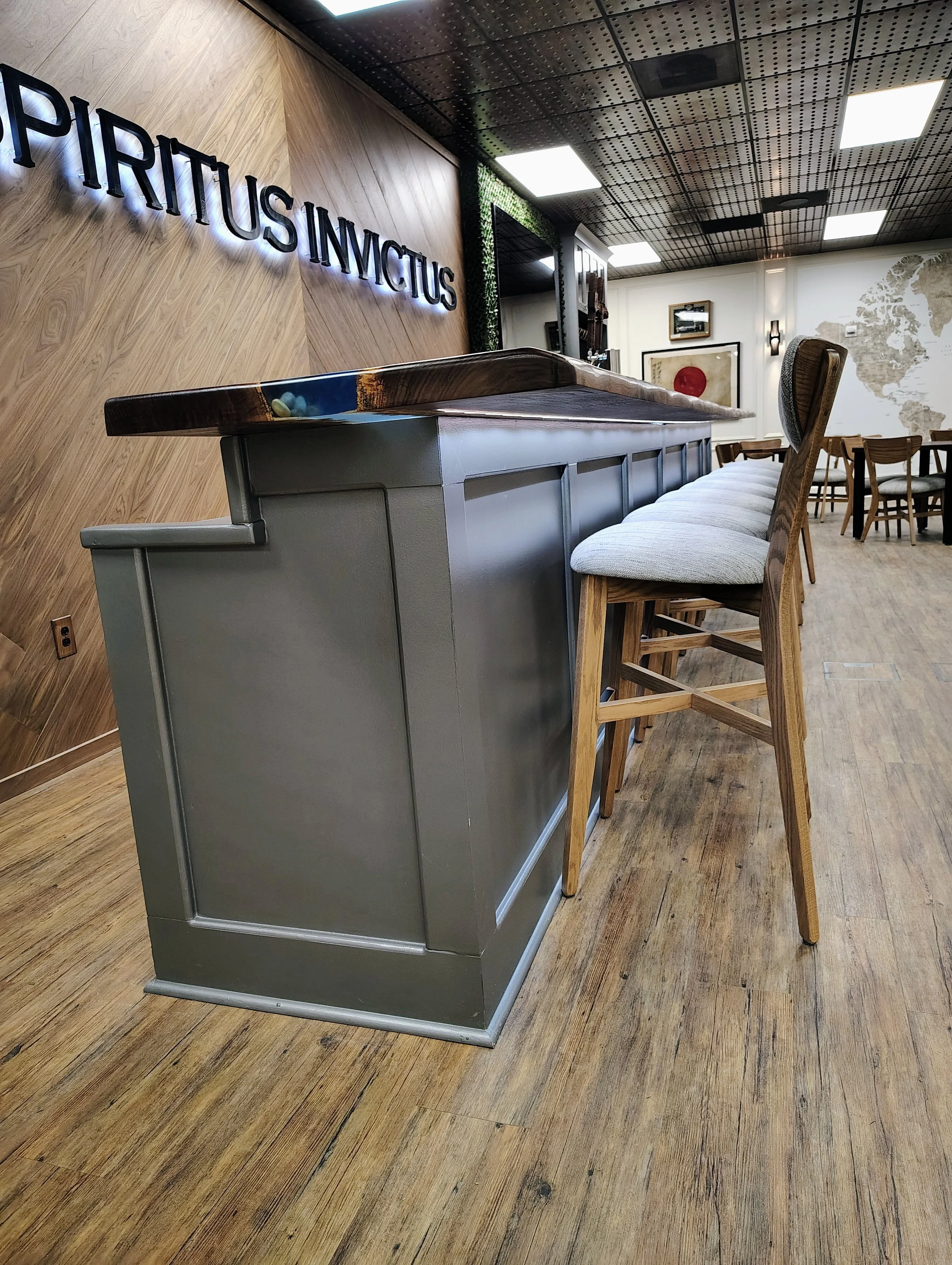Interior of a modern office or lounge space with a wooden accent wall, a bar-height table with a wood and metal design, and multiple chairs. There is a large wall map on the back wall and framed artwork. The ceiling has a black grid pattern with rece