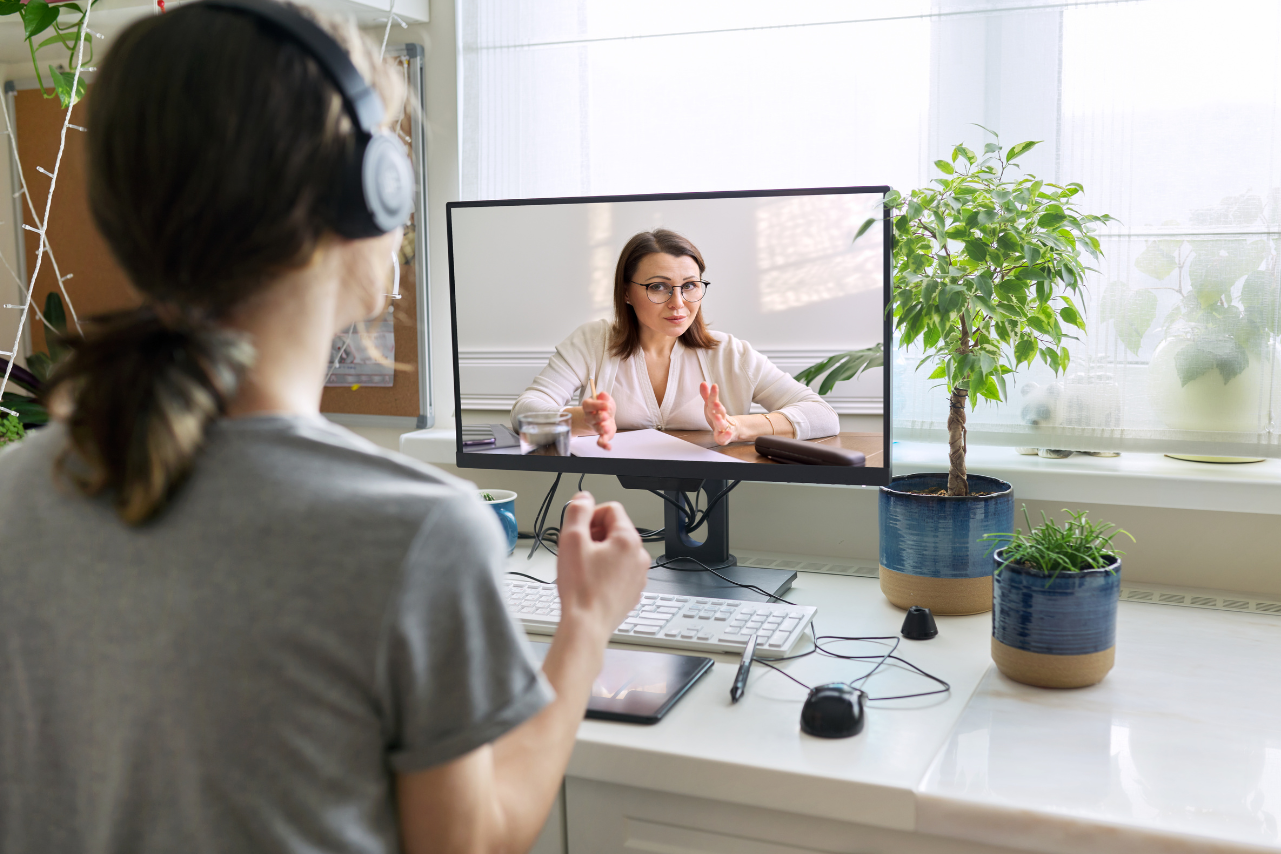 Woman participating in a video call with a woman on the computer screen, sitting at a desk with plants and a window in the background.