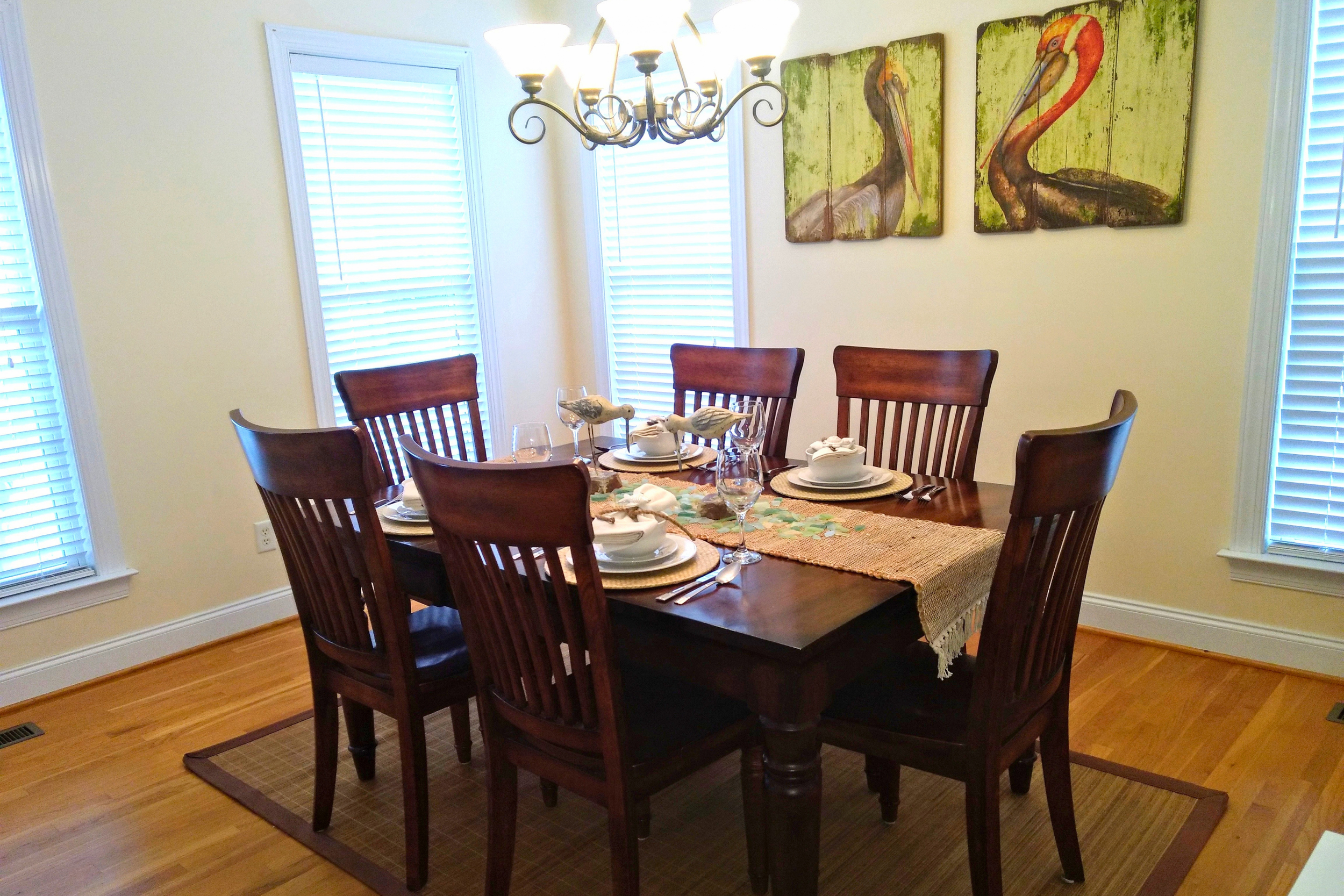 A dining room with a wooden table set for six people, including plates, glasses, and decorative bird figurines. The room has two large windows with white blinds, a small rug underneath the table, and a set of three canvas paintings of herons hanging on the wall. A chandelier with multiple lights hangs above the table.