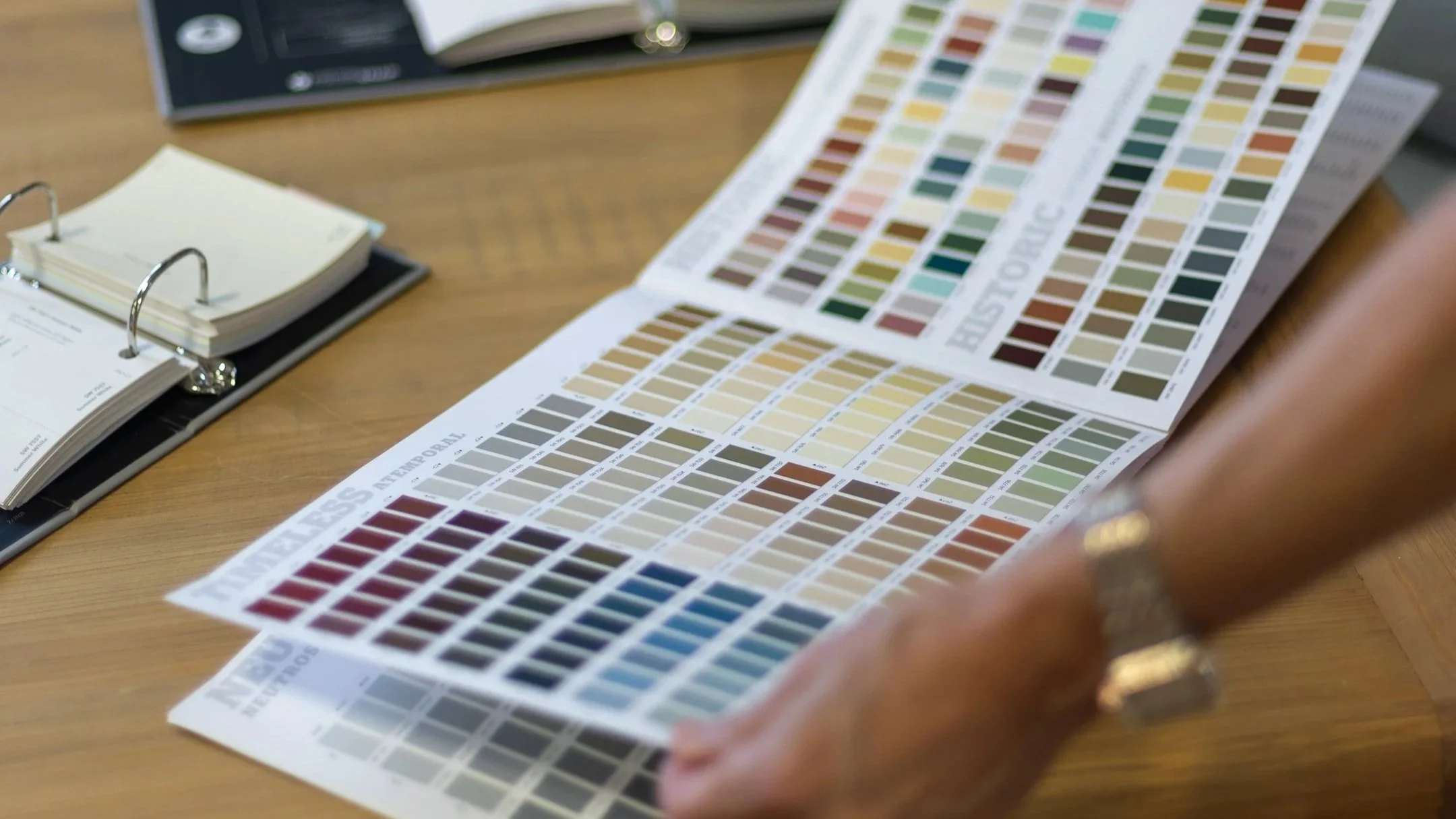 Close-up of a person holding a color sample chart with various shades arranged in rows and columns on a desk with notebooks and a phone nearby.
