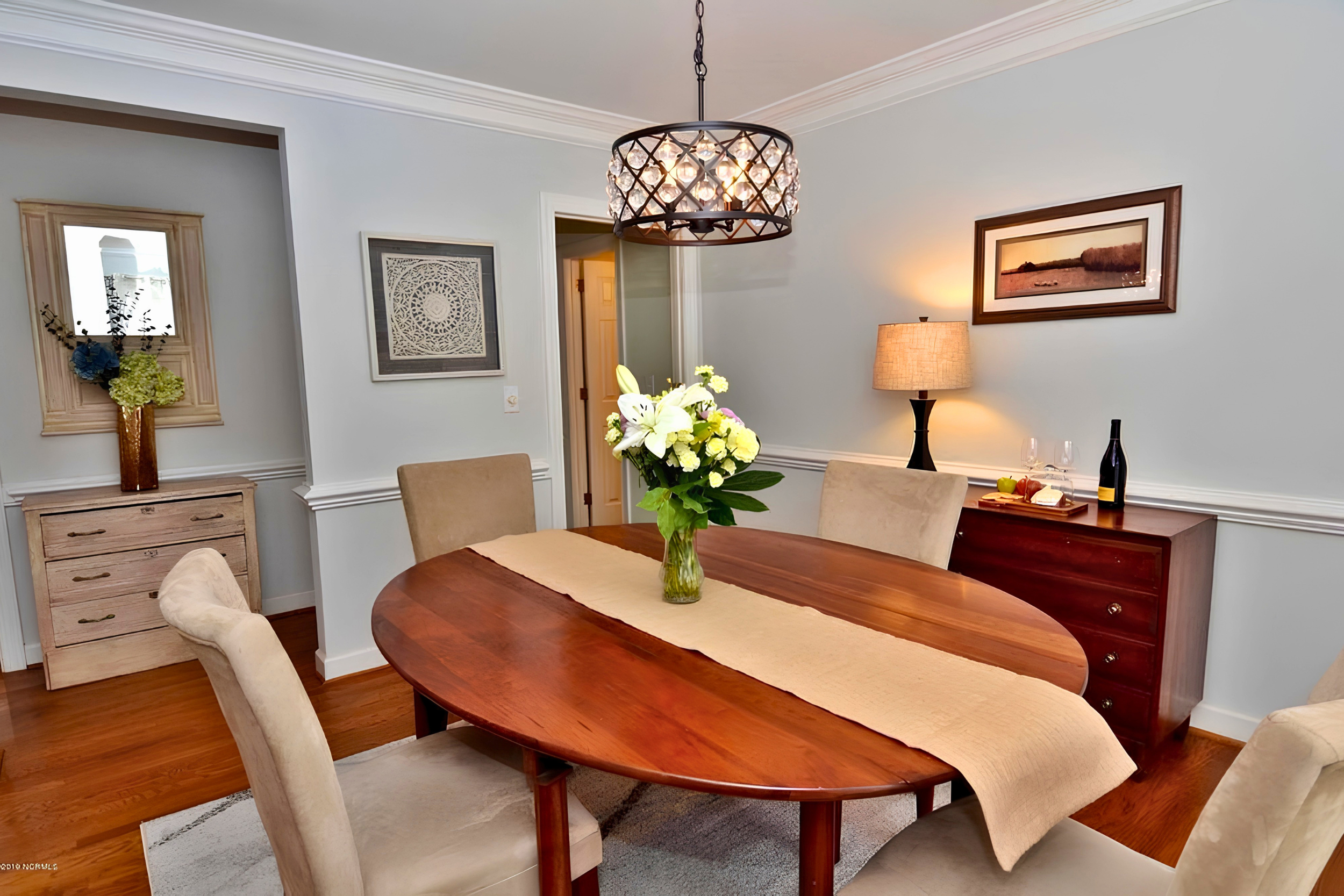 A dining room with a wooden oval table, beige upholstered chairs, a vase with white and yellow flowers, a chandelier hanging from the ceiling, framed artworks on the walls, a lamp, and a sideboard with wine and cheese.