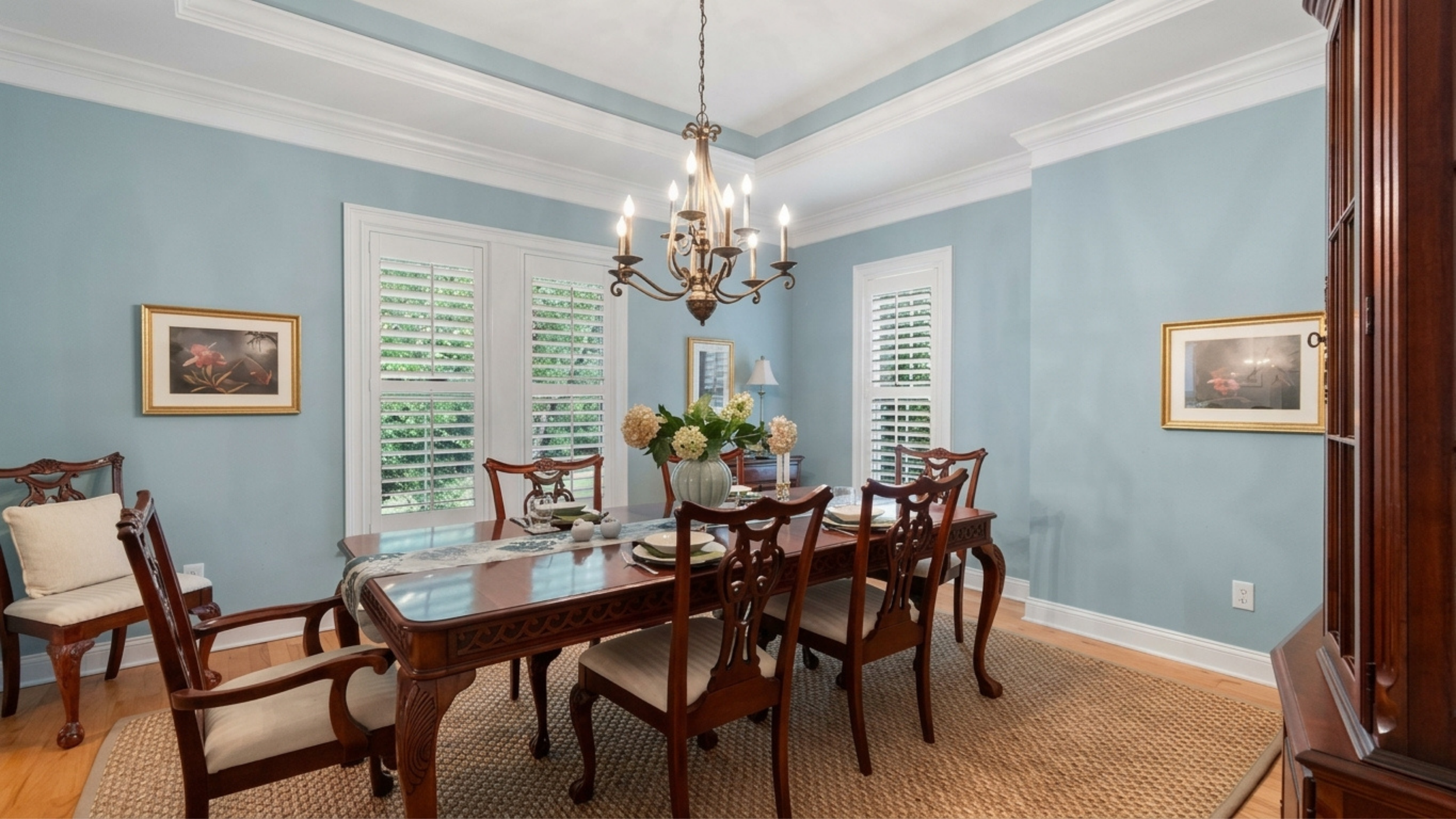 A dining room with light blue walls, white trim, and large windows with white shutters. A chandelier hangs over a wooden dining table set with placemats, napkins, and a floral centerpiece. There are wooden chairs with cushioned seats, a wooden sideboard, and framed floral artwork on the walls.