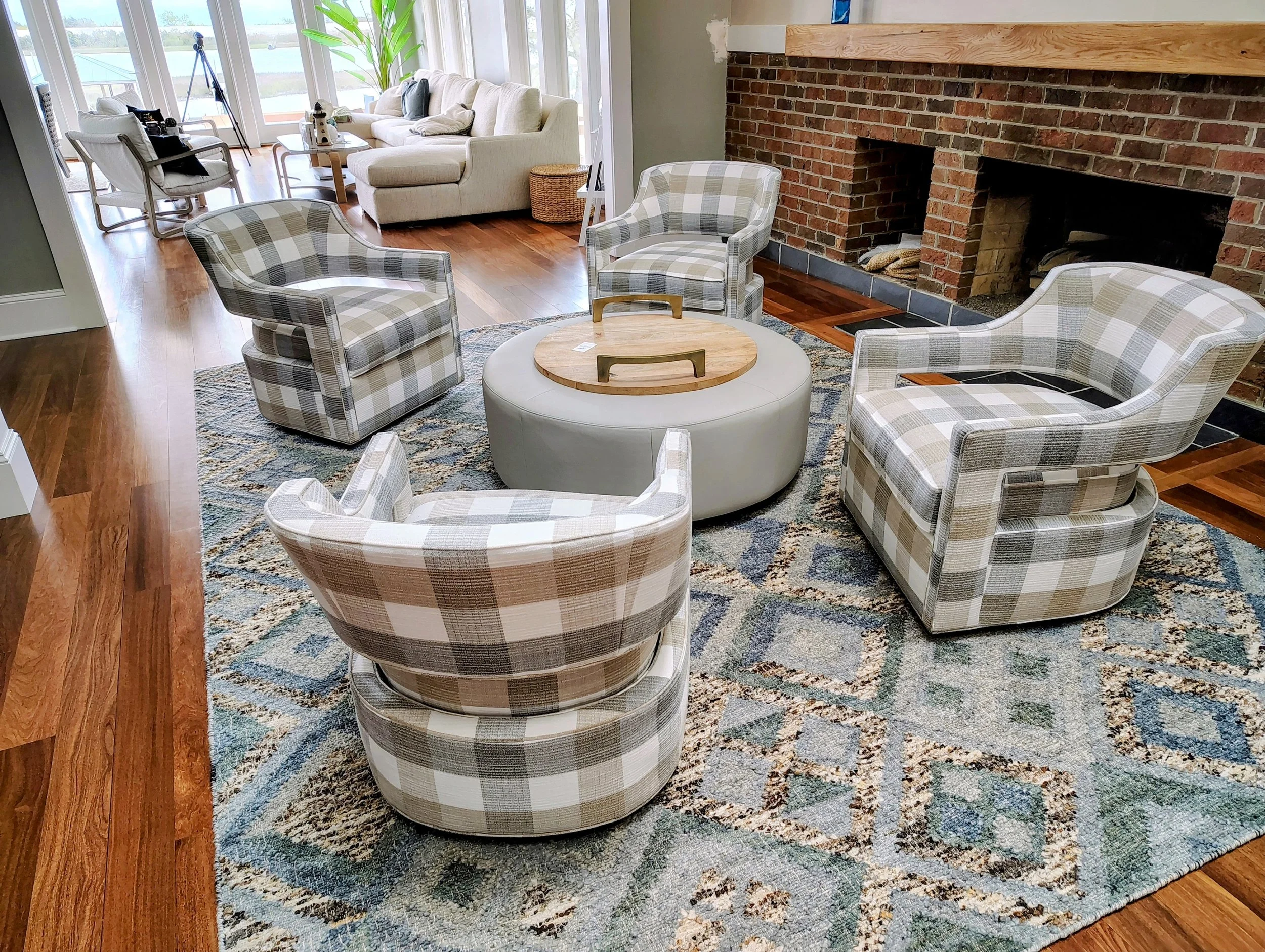 A living room with a brick fireplace, plaid armchairs, a round ottoman with a wooden tray, a patterned rug, and a white sofa near large windows with view of trees and water outside.