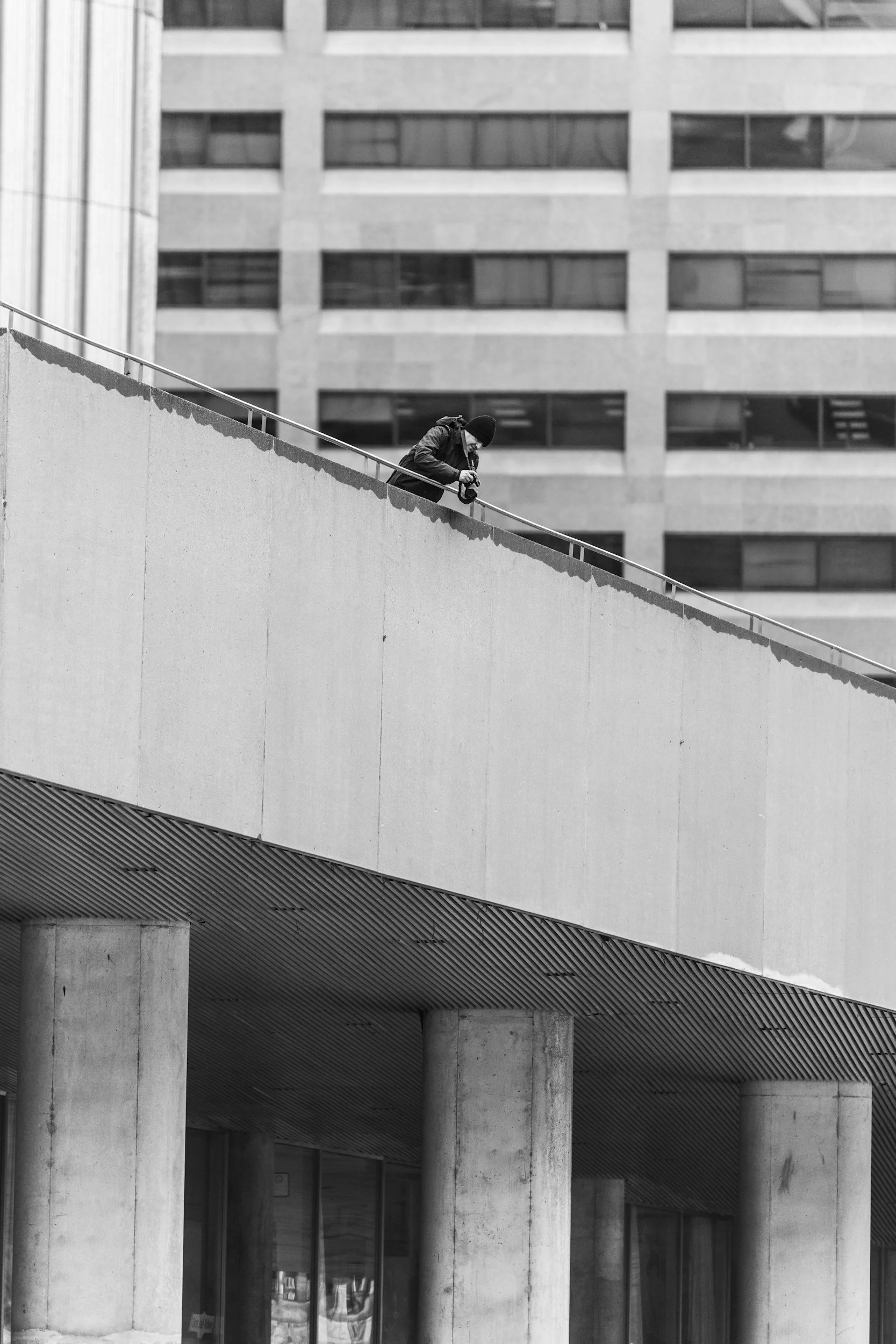 Black and white photo of a person leaning over a city building's edge with a camera, capturing urban architecture.