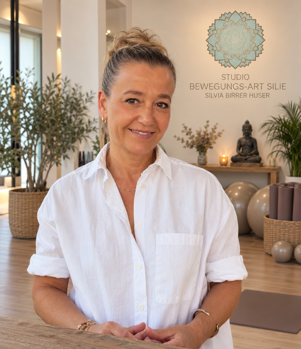 Eine lachende Frau mit blonden Haaren in einem weißen Hemd in einem Yoga-Studio, mit Buddha-Statuen, Pflanzen und Yogamatten im Hintergrund.