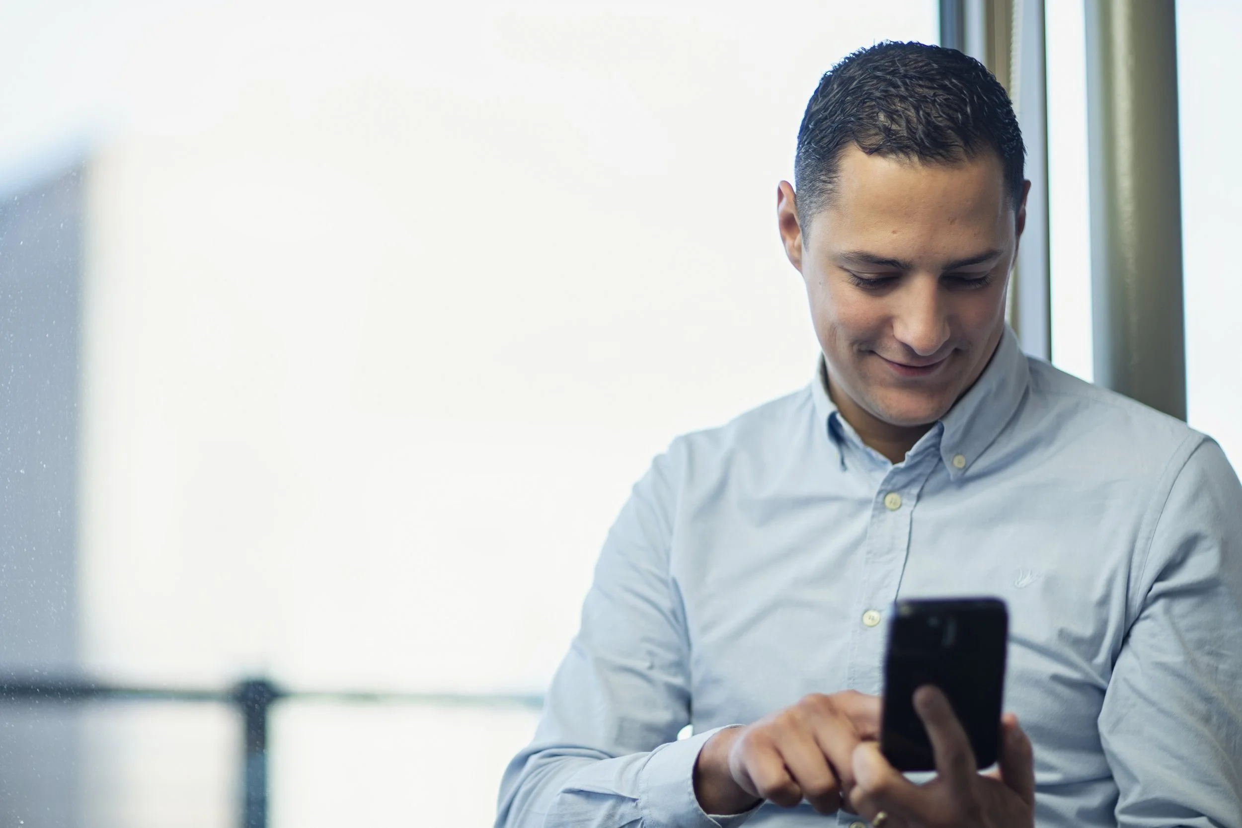 A young man in a light blue button-up shirt looking at and using a smartphone, standing near a window with natural light.