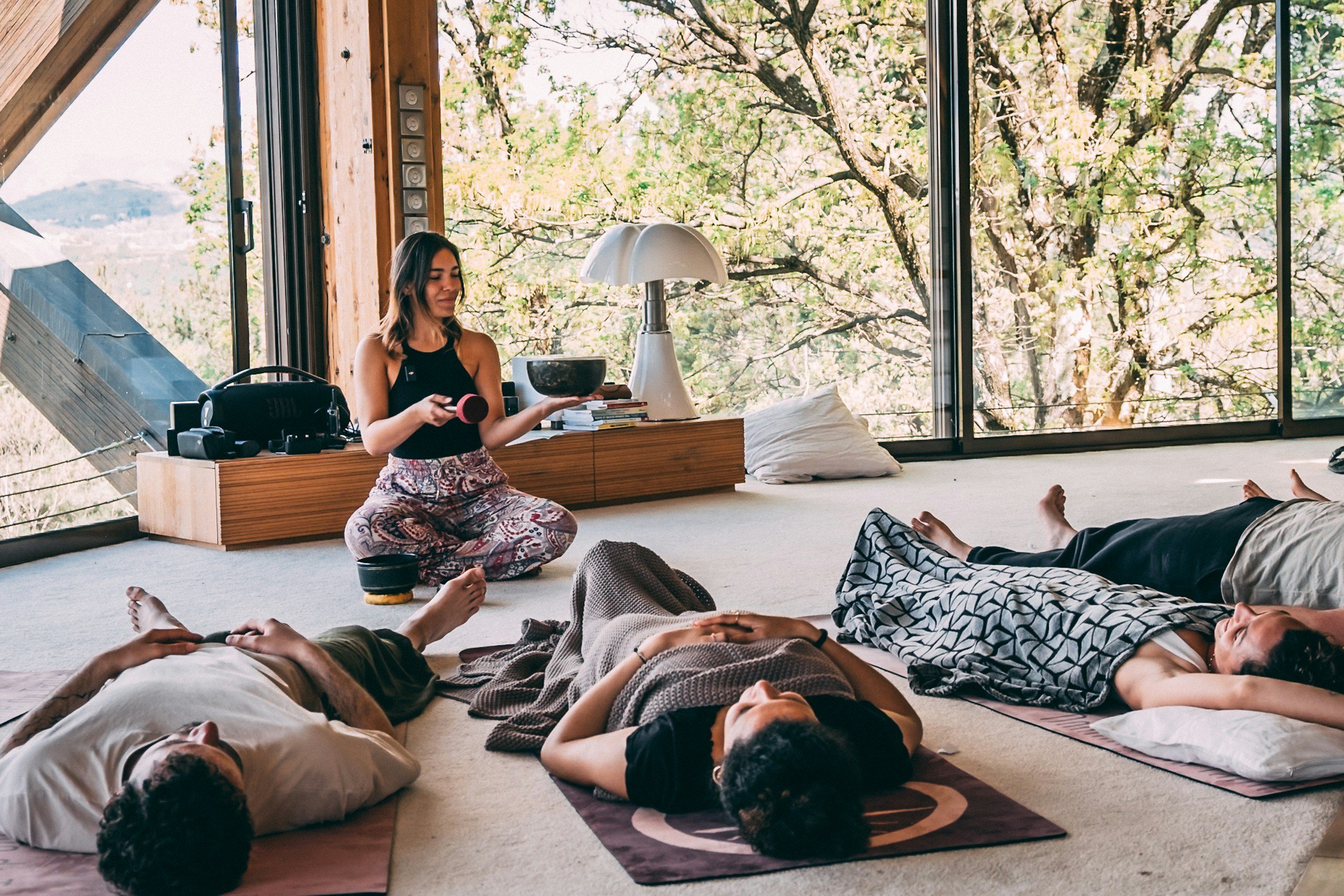 Group of people lying on mats during a yoga or meditation session in a room with large windows showing trees outside. One person sits up holding a singing bowl, surrounded by various objects and books.