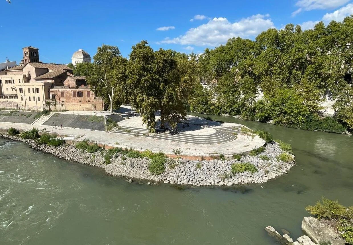 A scenic riverside view with a paved area on a small peninsula, trees lining the background, and historic buildings with domed roofs in the distance.