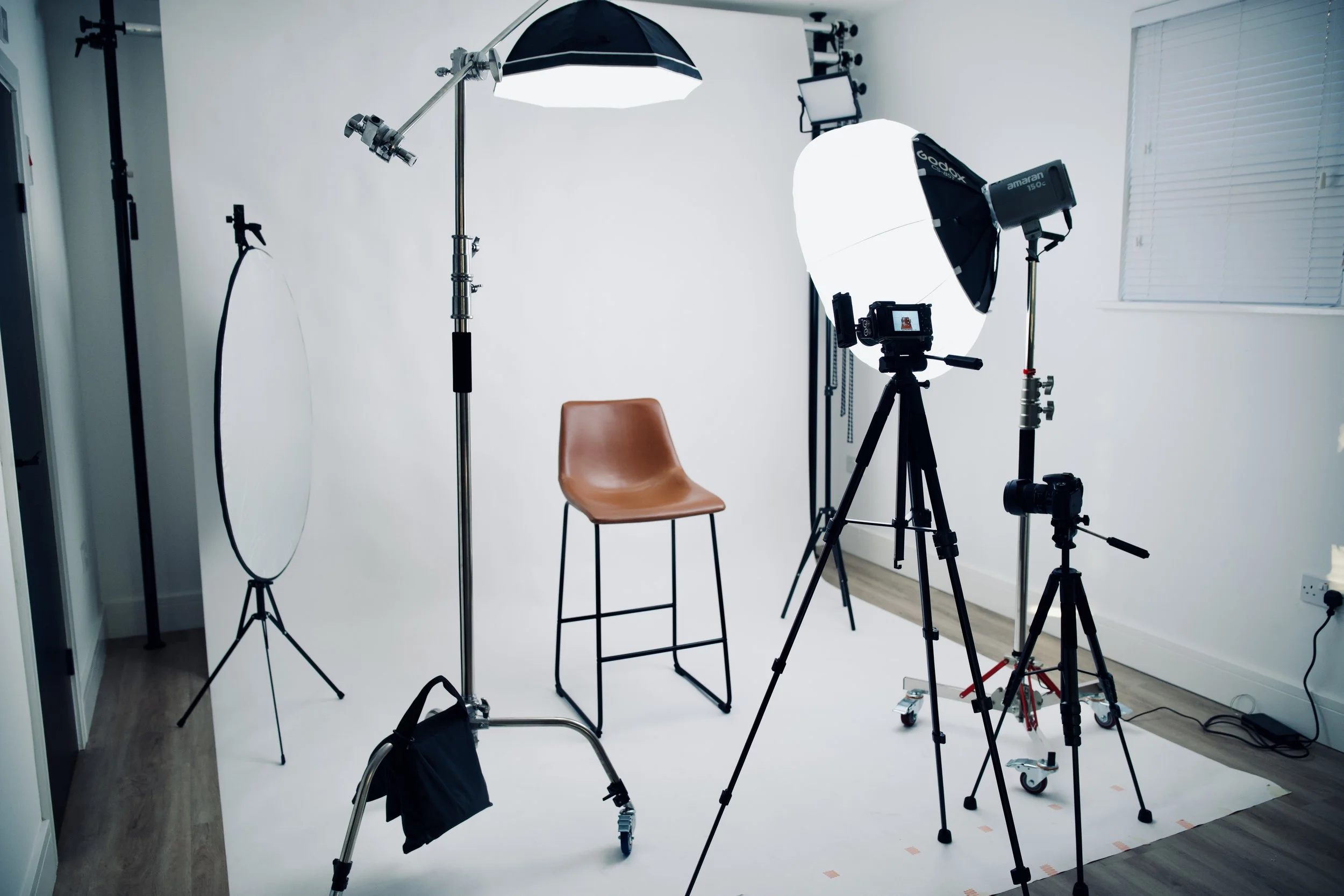 Photography studio setup with a brown chair, professional lighting equipment, cameras, and a white backdrop