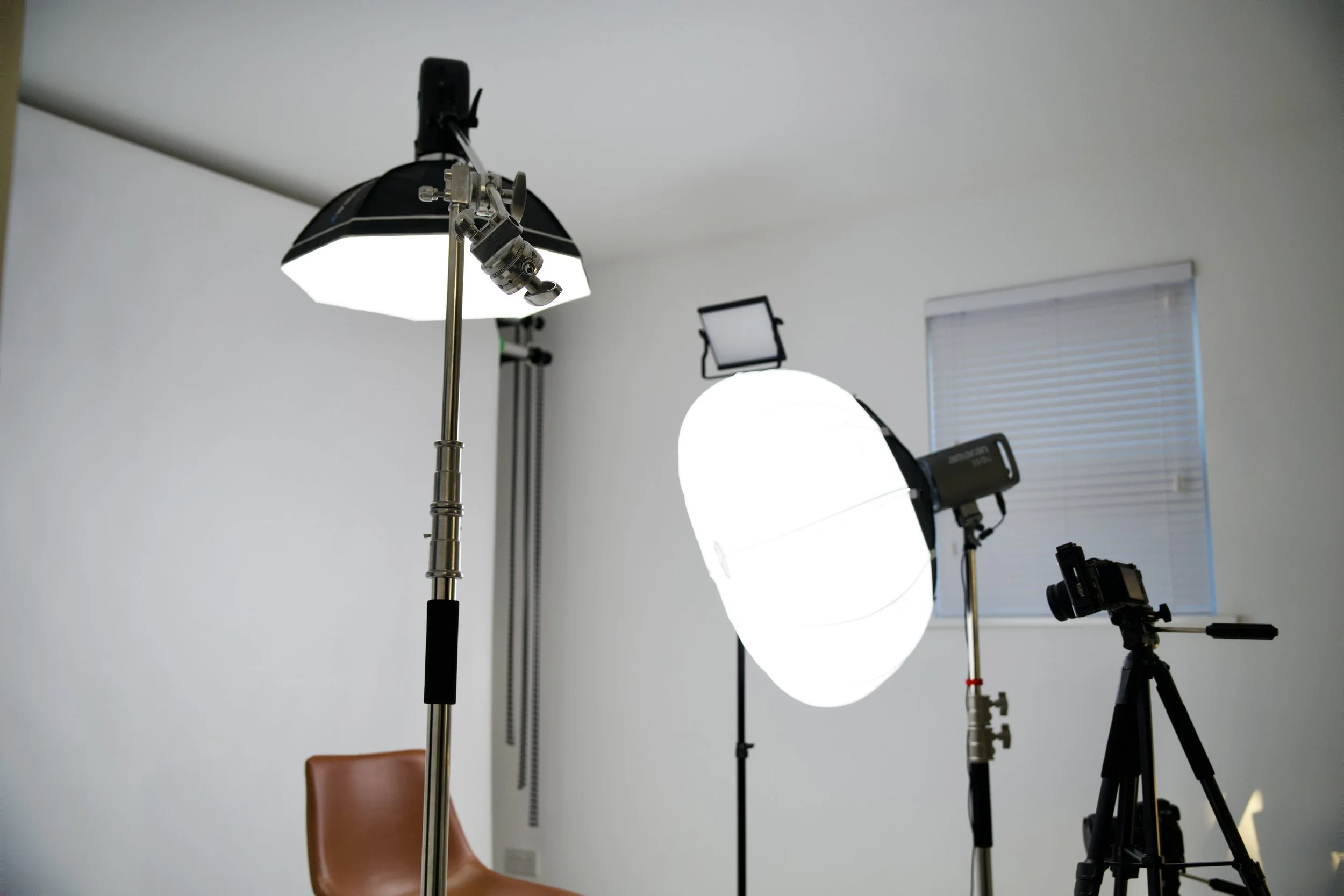 Photography studio setup with three professional light sources, a camera on a tripod, and a brown chair against a white wall with a window.