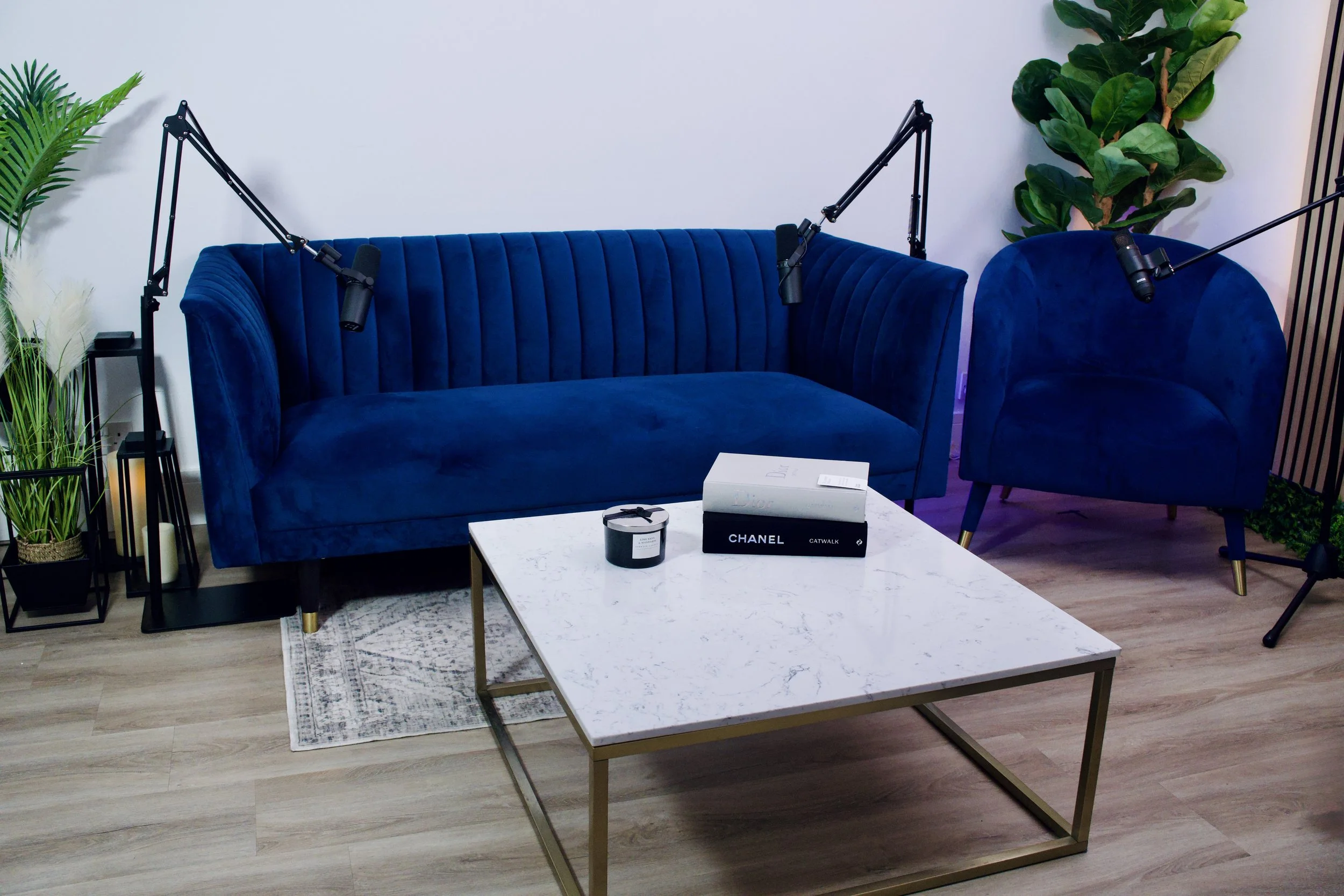 A modern living room with a blue velvet sofa and matching armchair, a white marble coffee table, and decorative plants. Two microphones are positioned near the sofa and armchair, suggesting a podcast or recording setup.