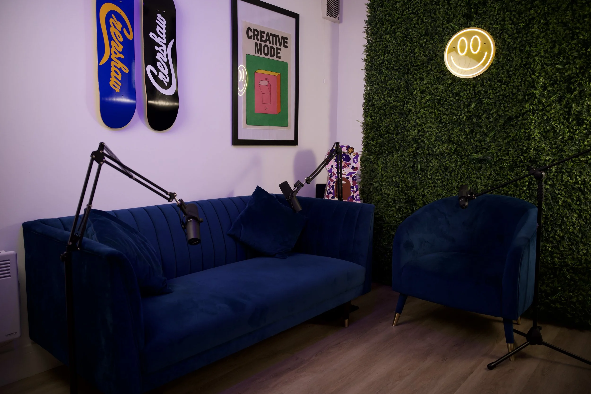 A cozy room with a dark blue velvet sofa and matching armchair, framed pictures on the wall, a green faux green wall with a smiley face neon sign, a guitar in the corner, and microphone stands.
