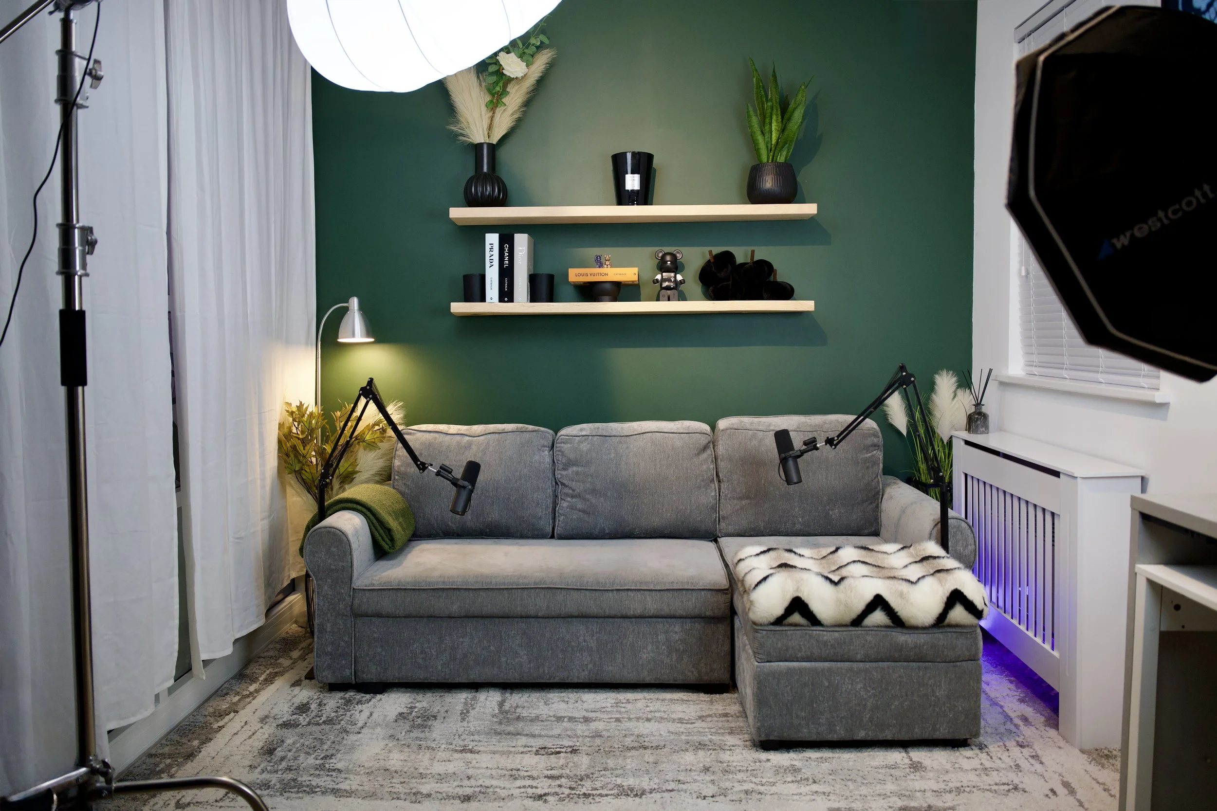 Living room with gray sofa against a green wall, two black microphone arms attached to the sofa, white curtains on the left, a white side table with plants on the right, and two shelves with decorative items and books above the sofa. Studio lighting
