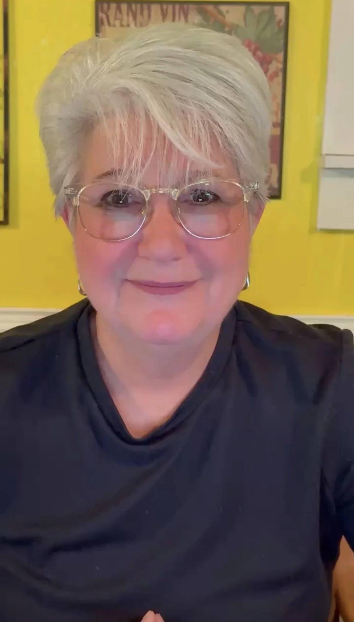 A smiling woman with short gray hair, glasses, and earrings, sitting in front of a bright yellow wall with framed artwork.