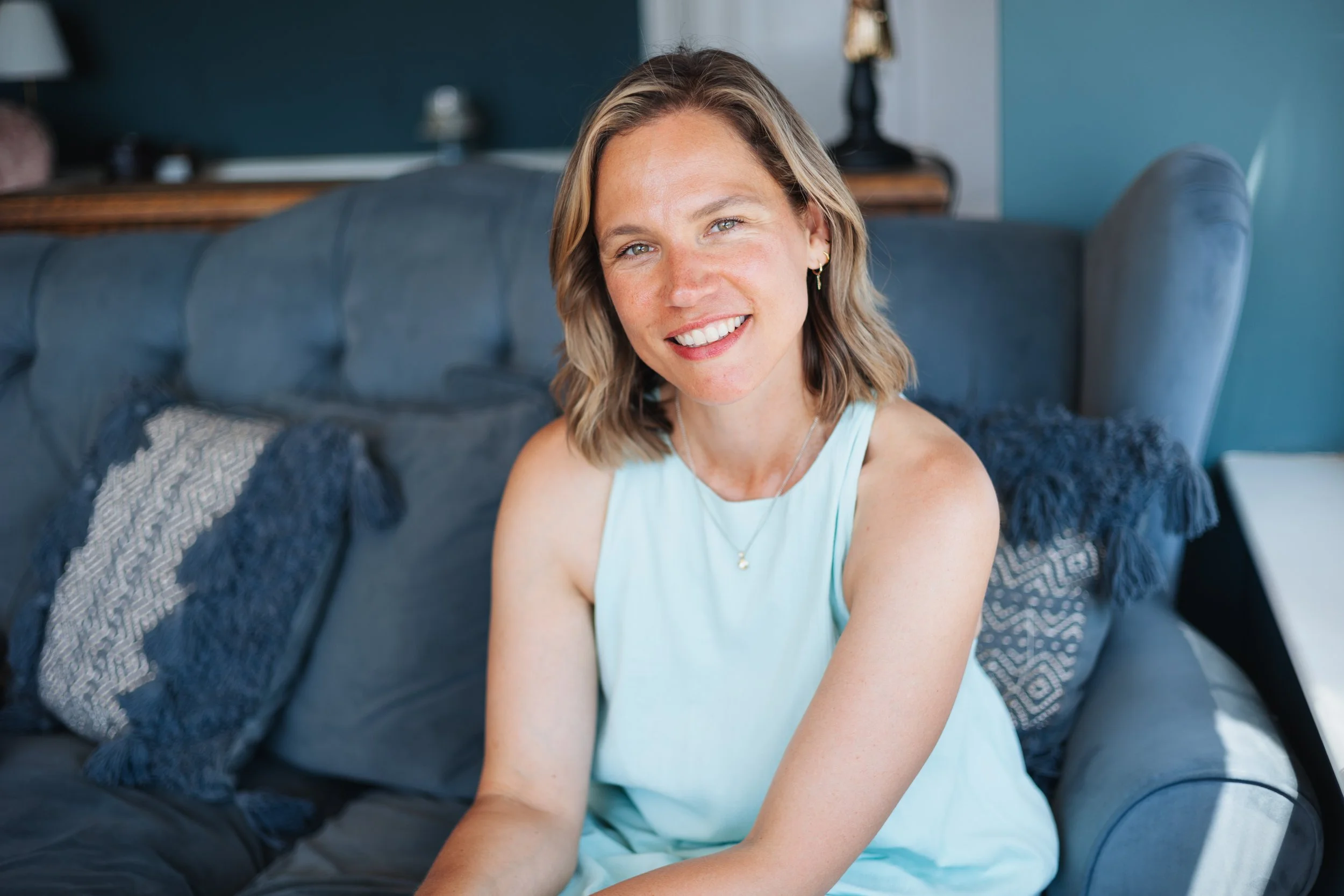 Louise with shoulder-length blonde hair and earrings, wearing a sleeveless light blue top, sitting on a dark blue couch with patterned and fringed pillows, smiling at the camera in a living room.