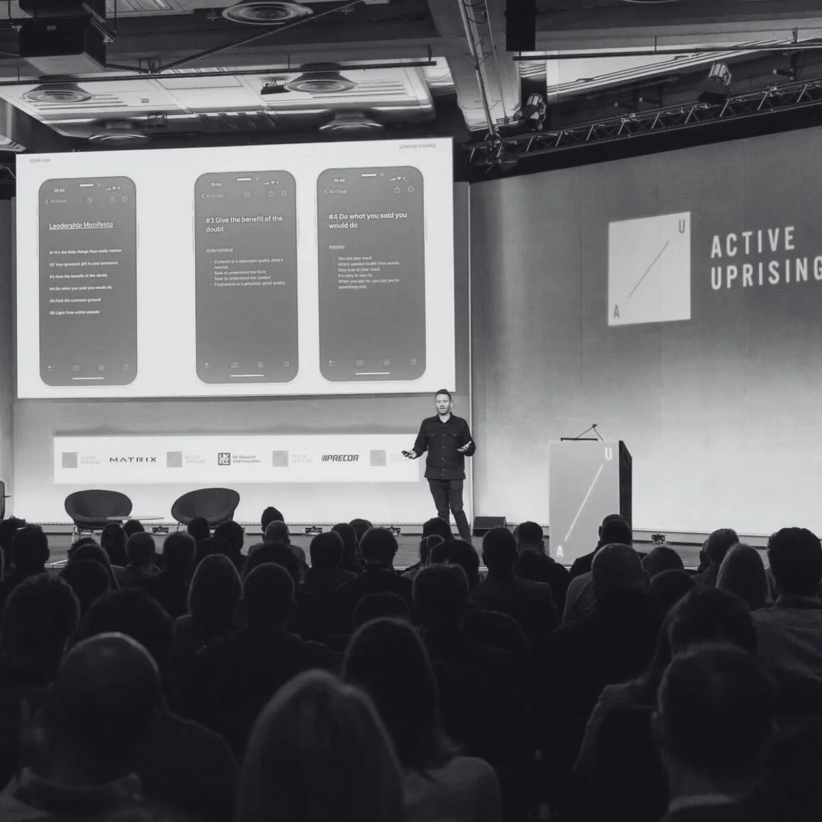A man giving a presentation on a stage to a seated audience. The large screen behind him displays three smartphone screens with text, titled 'Leadership Manifesto' and numbered points. To the right of the stage, a large screen shows a diagram labeled 'ACTIVE UPRISING' with a square divided into four parts labeled 'U' and 'A'. There are three chairs on stage, and branding logos are visible below the screen.