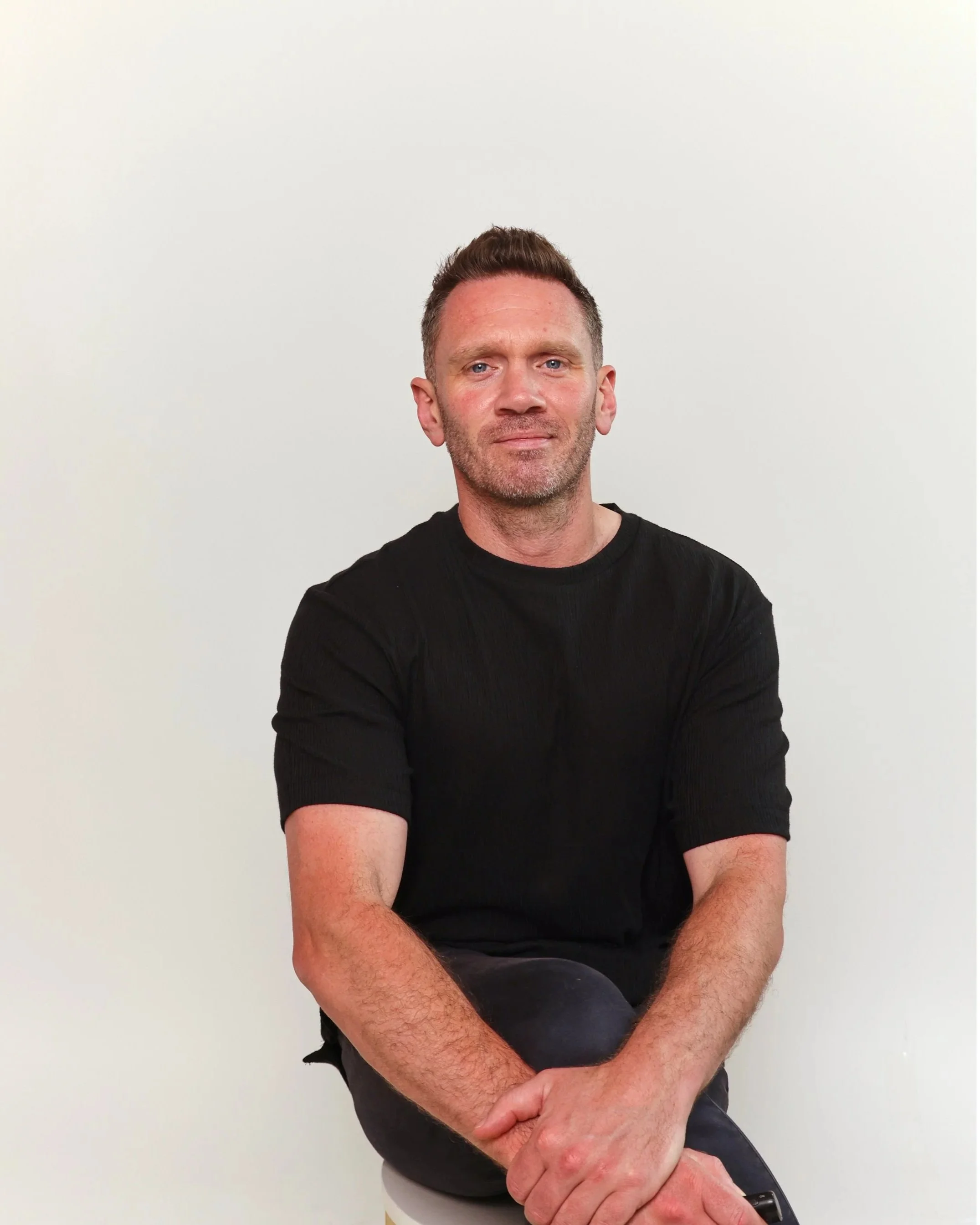 A man with short brown hair, blue eyes, and light facial hair, wearing a black T-shirt, sitting against a plain off-white wall.