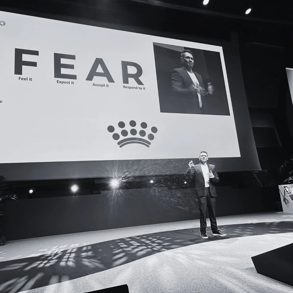 A man in a suit giving a presentation on stage with a large screen behind him displaying the word 'FEAR' and related words 'Feel it,' 'Expect it,' 'Accept it,' 'Respond to it,' along with a photo of himself and a crown icon.