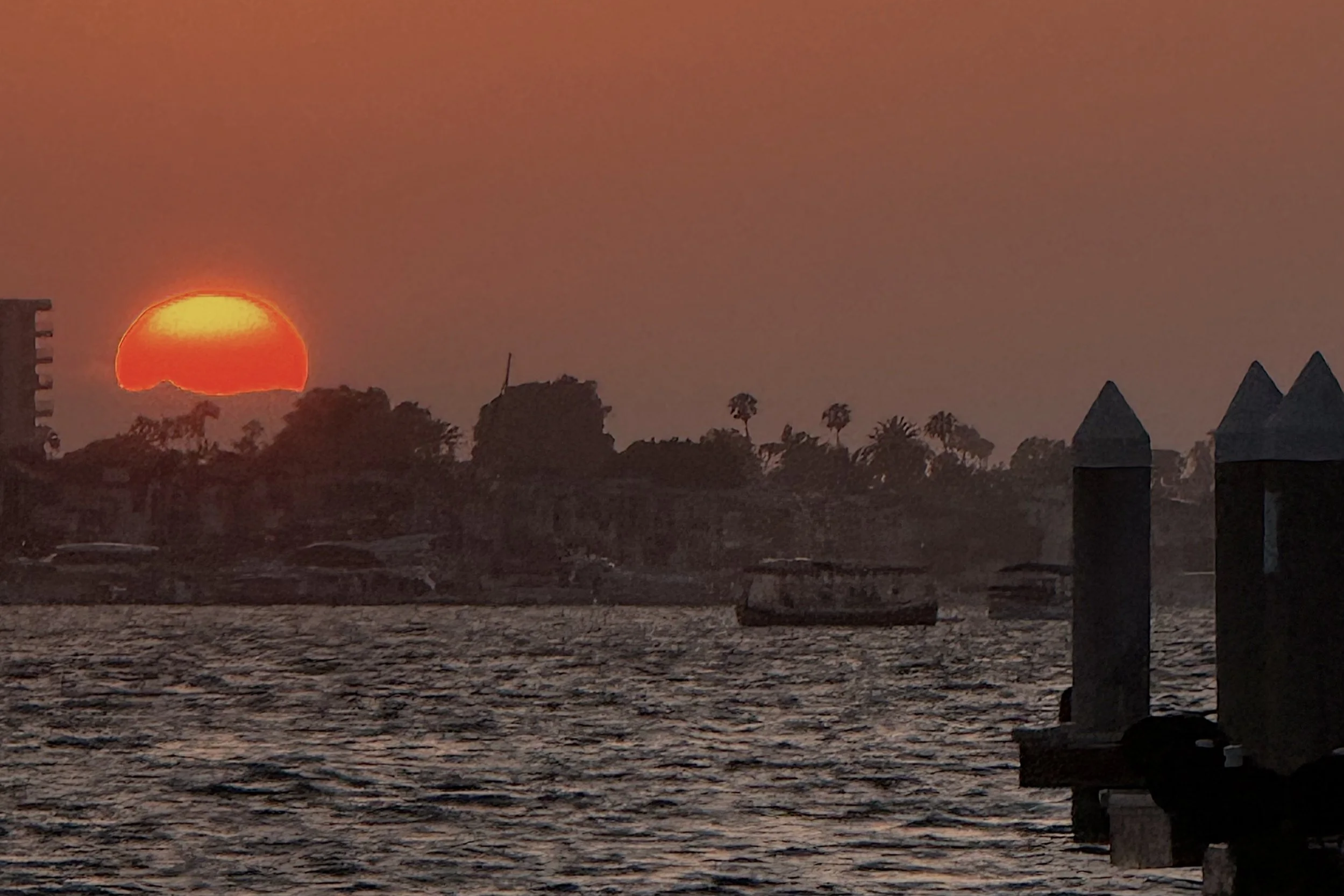 Sunset over water with visible cityscape and silhouettes of trees and buildings.