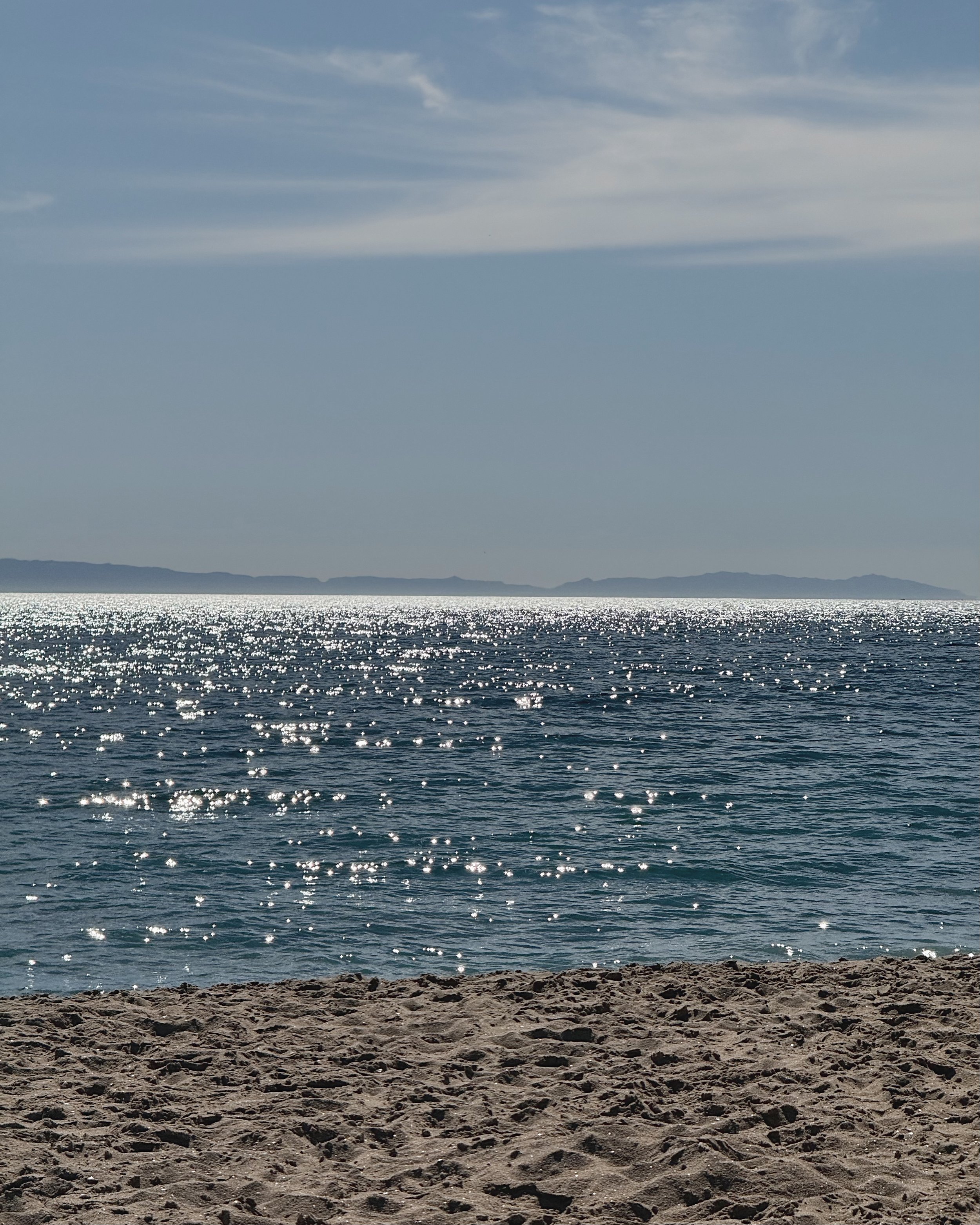 A sandy beach with small rocks in the foreground, sparkling water in the middle, and distant land or mountains under a partly cloudy sky in the background.