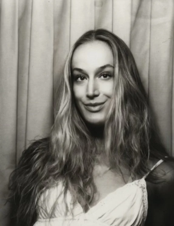 Black and white photo of a young woman with long wavy hair, smiling slightly, standing in front of curtains.