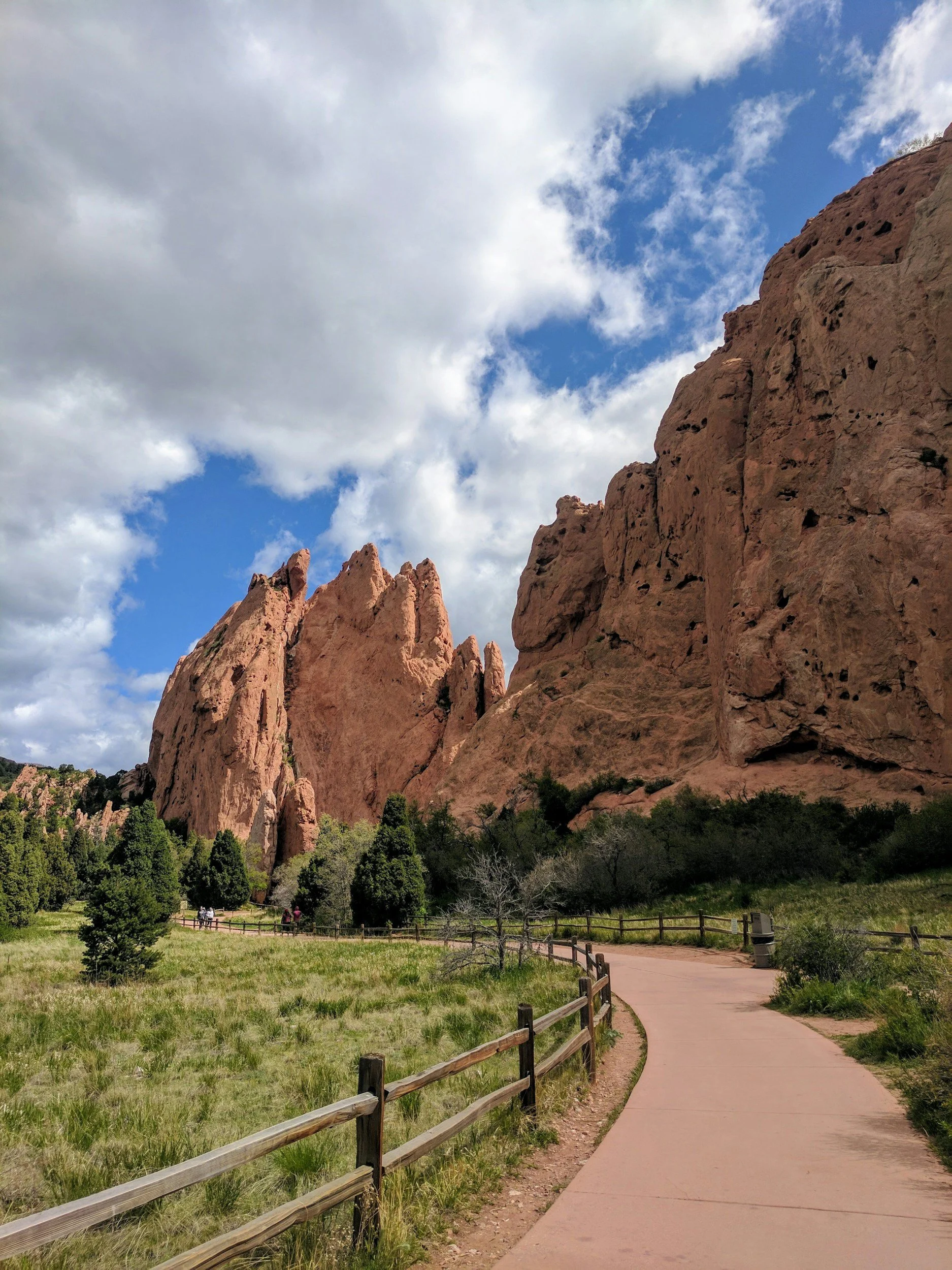 A scenic view of red rock formations with a curved walking path, wooden fence, green trees, and a partly cloudy blue sky.