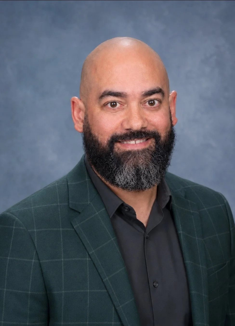 Headshot of a man with a shaved head and a beard, wearing a dark shirt and green plaid blazer, smiling against a blue background.