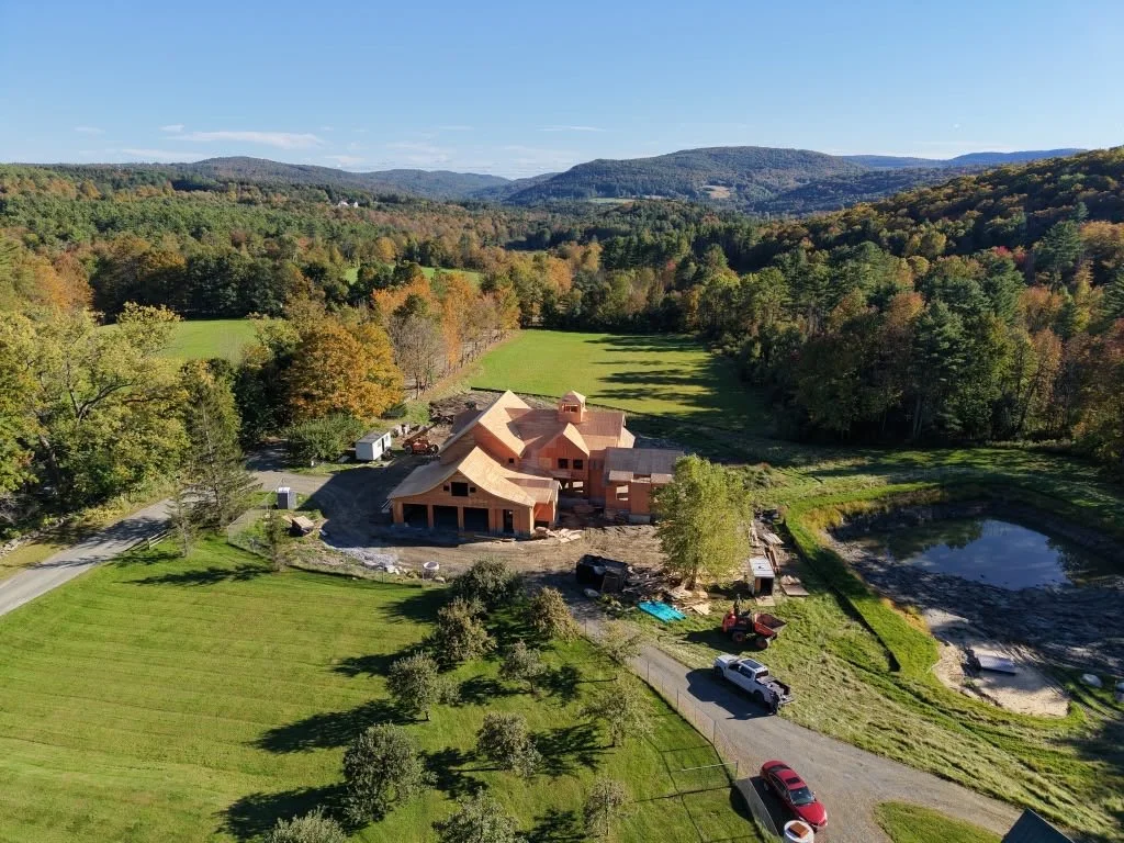 A house under construction in a rural area surrounded by green fields, trees, and hills with a small pond nearby.