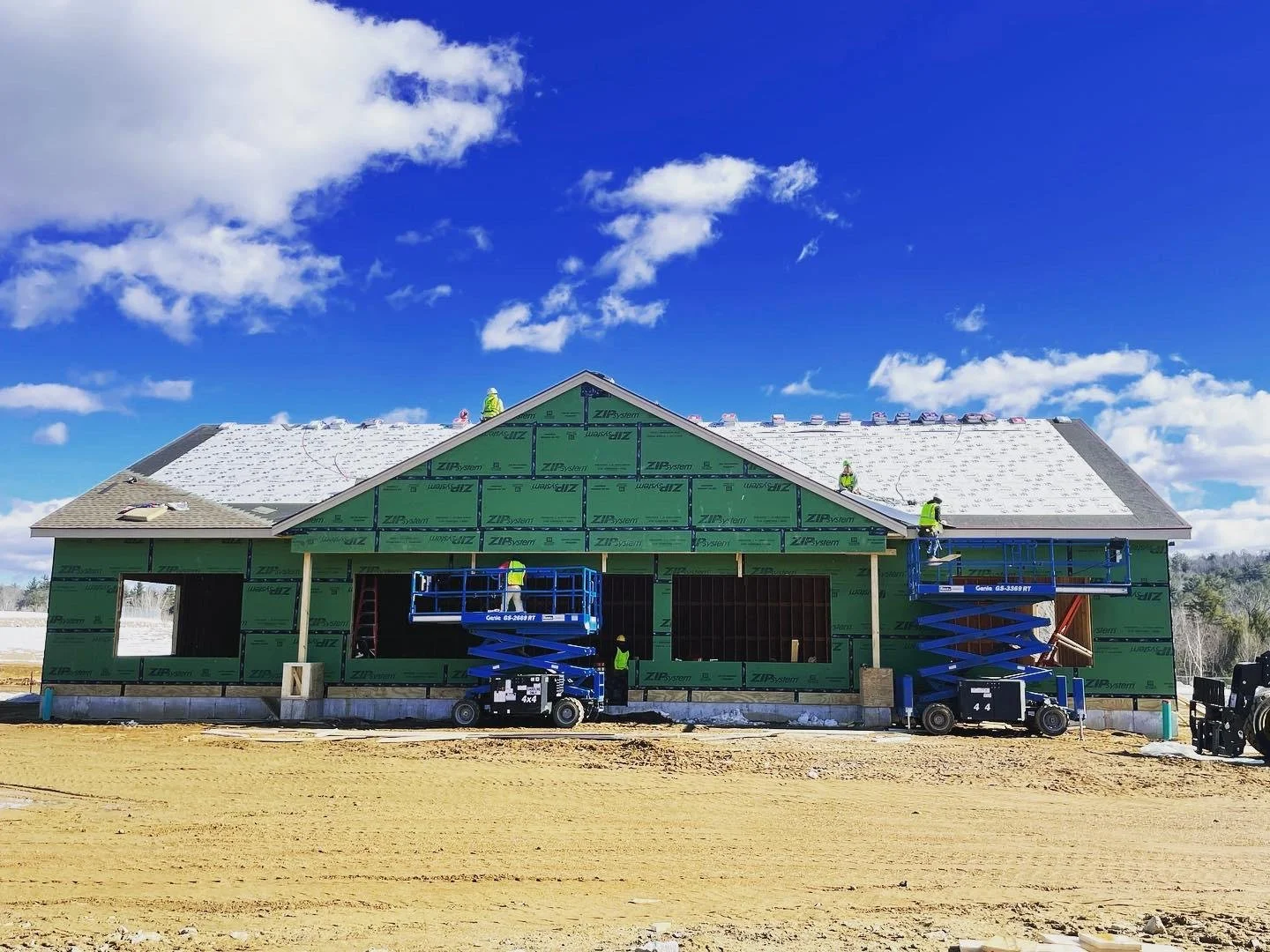 Construction workers installing roofing on a house under a partly cloudy sky. The house is wrapped in green sheathing material, with some sections of the roof already covered with shingles.