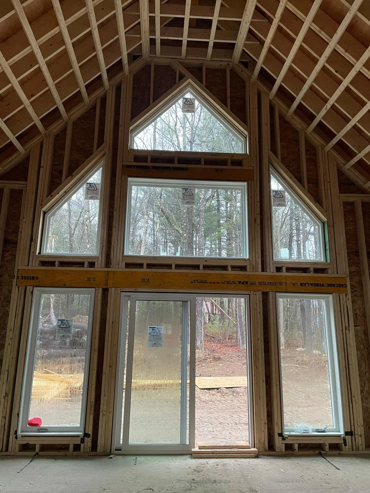 Interior view of a house under construction with wooden framing and large multiple-pane windows, overlooking a wooded outdoor area.