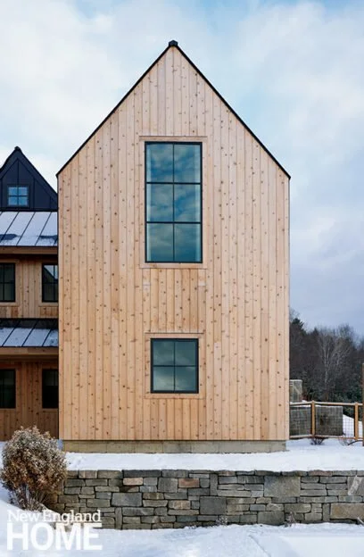 A modern house with a wooden exterior and large rectangular windows, situated on a stone foundation, with snow on the ground.