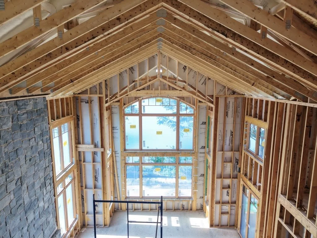 Interior of a house under construction with exposed wooden framing, large front windows, and a stone accent wall on the left.