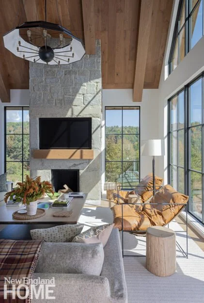 Living room with large windows, stone fireplace, wooden ceiling beams, leather chairs, sofa, and a fireplace with a wooden mantel.