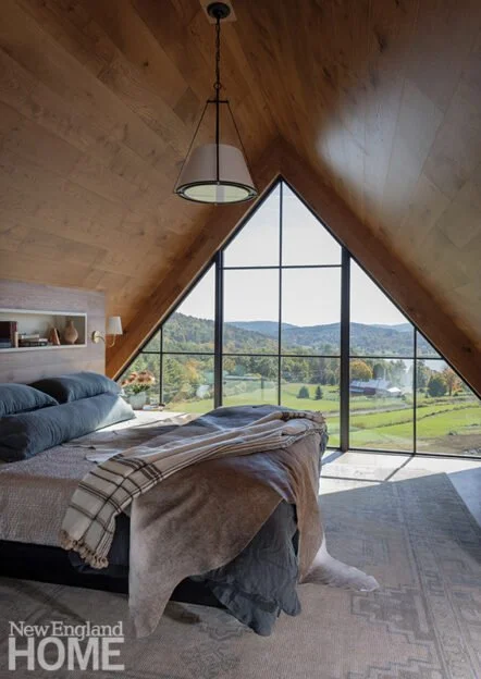 A bedroom with an A-frame ceiling and large triangular window showing a scenic view of green fields and trees. The room has a bed with a blanket and pillows, a hanging lamp, and a small shelf with decor items.