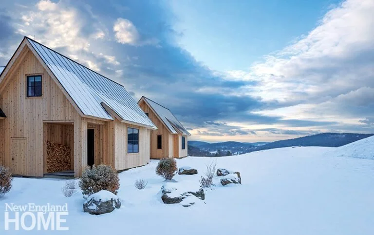 Two wooden house structures with metal roofs on a snowy landscape under a partly cloudy sky, with hills in the background.