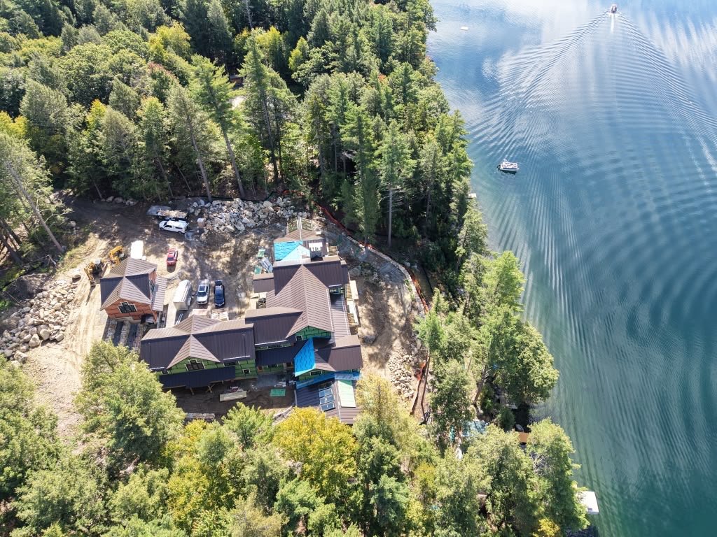 An aerial view of a lakeside property with multiple buildings and vehicles, surrounded by trees, alongside a large body of water with boats and ripples.