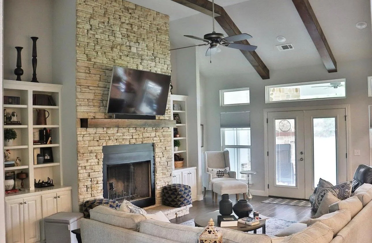 Living room with beige sofa, stone fireplace with wall-mounted TV, white built-in shelves, wooden ceiling beams, large windows, and a glass front door.