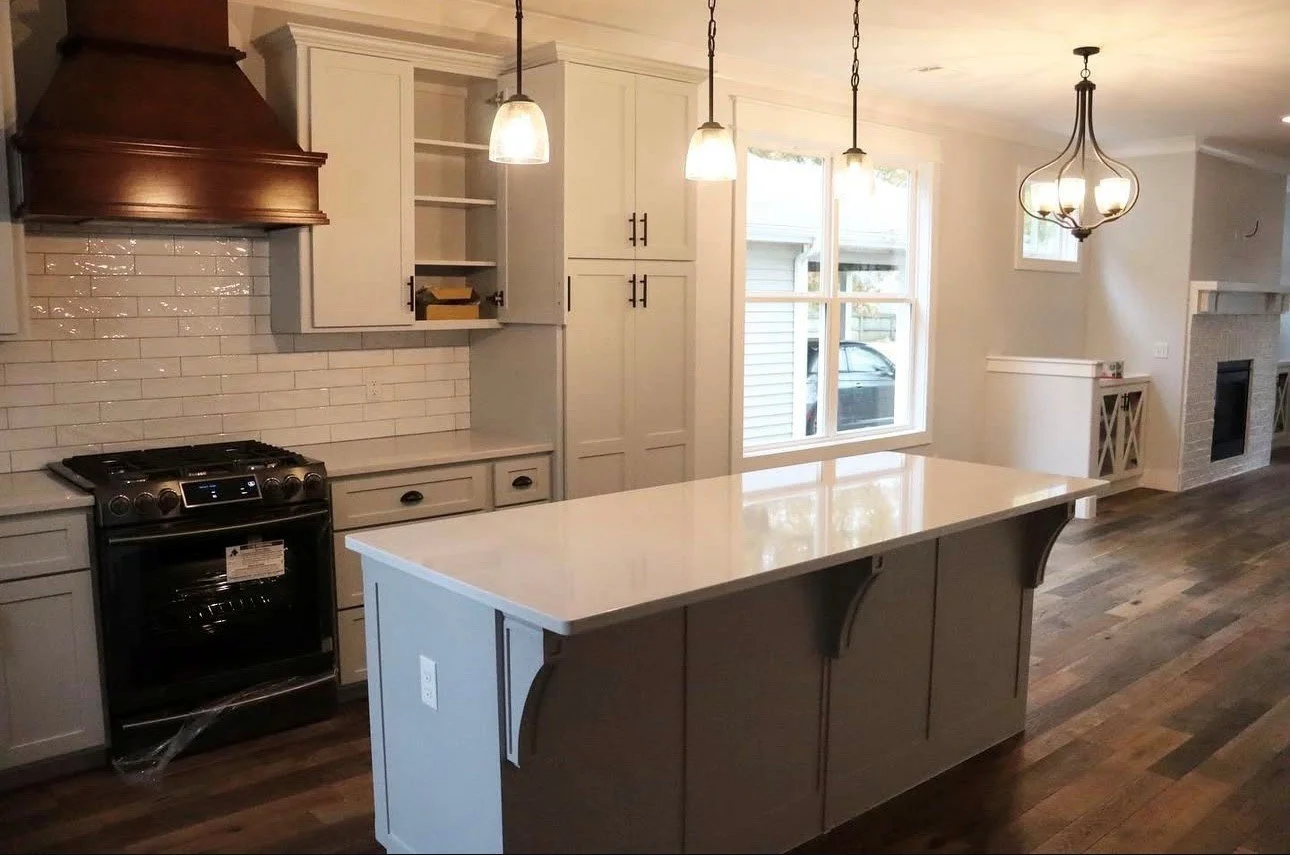 Modern kitchen with white cabinets, a central island with a white countertop, a black stove, pendant lights, and a large window overlooking the outside. A fireplace is visible in the background.